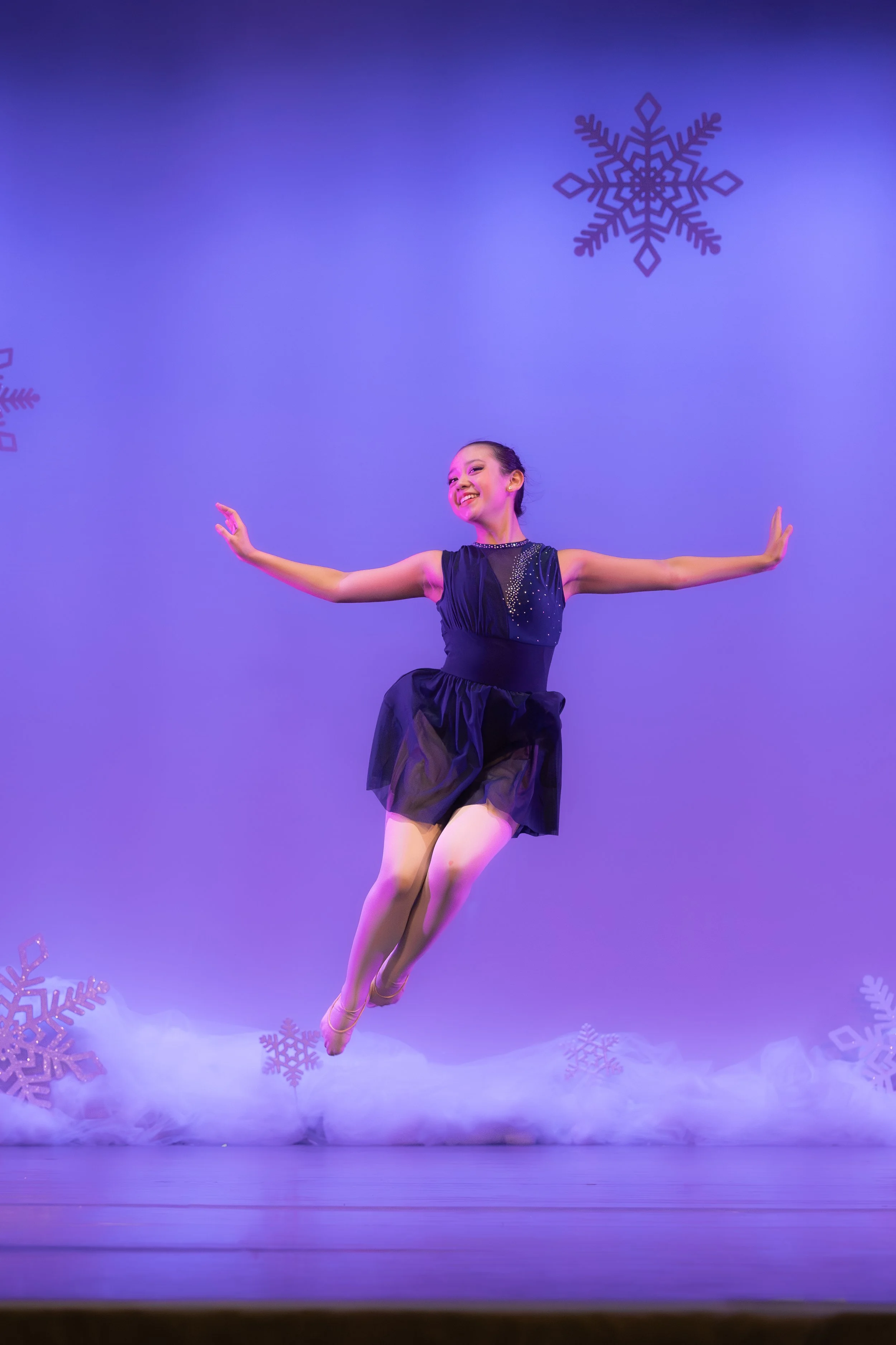 A young ballerina performing on stage, mid-air, with snowflake decorations and purple background.