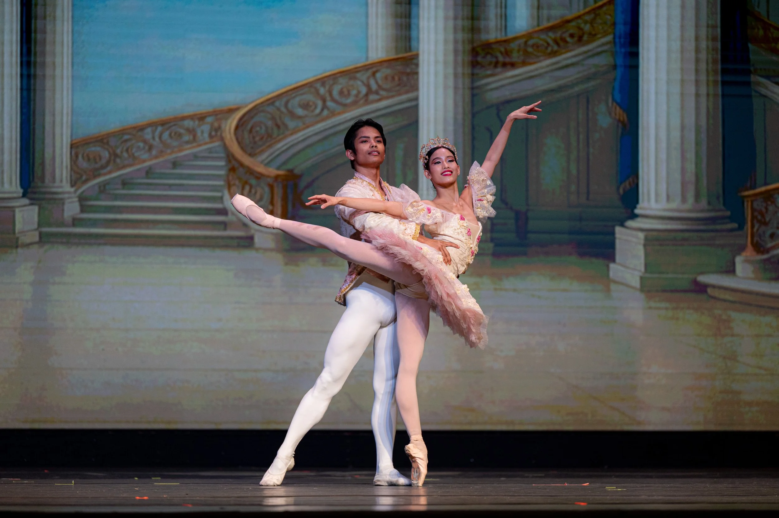 Two ballet dancers perform on stage with a painted backdrop of a grand staircase and columns. The male dancer supports the female dancer in an arabesque pose, with the female wearing a pink tutu and the male in a white costume.