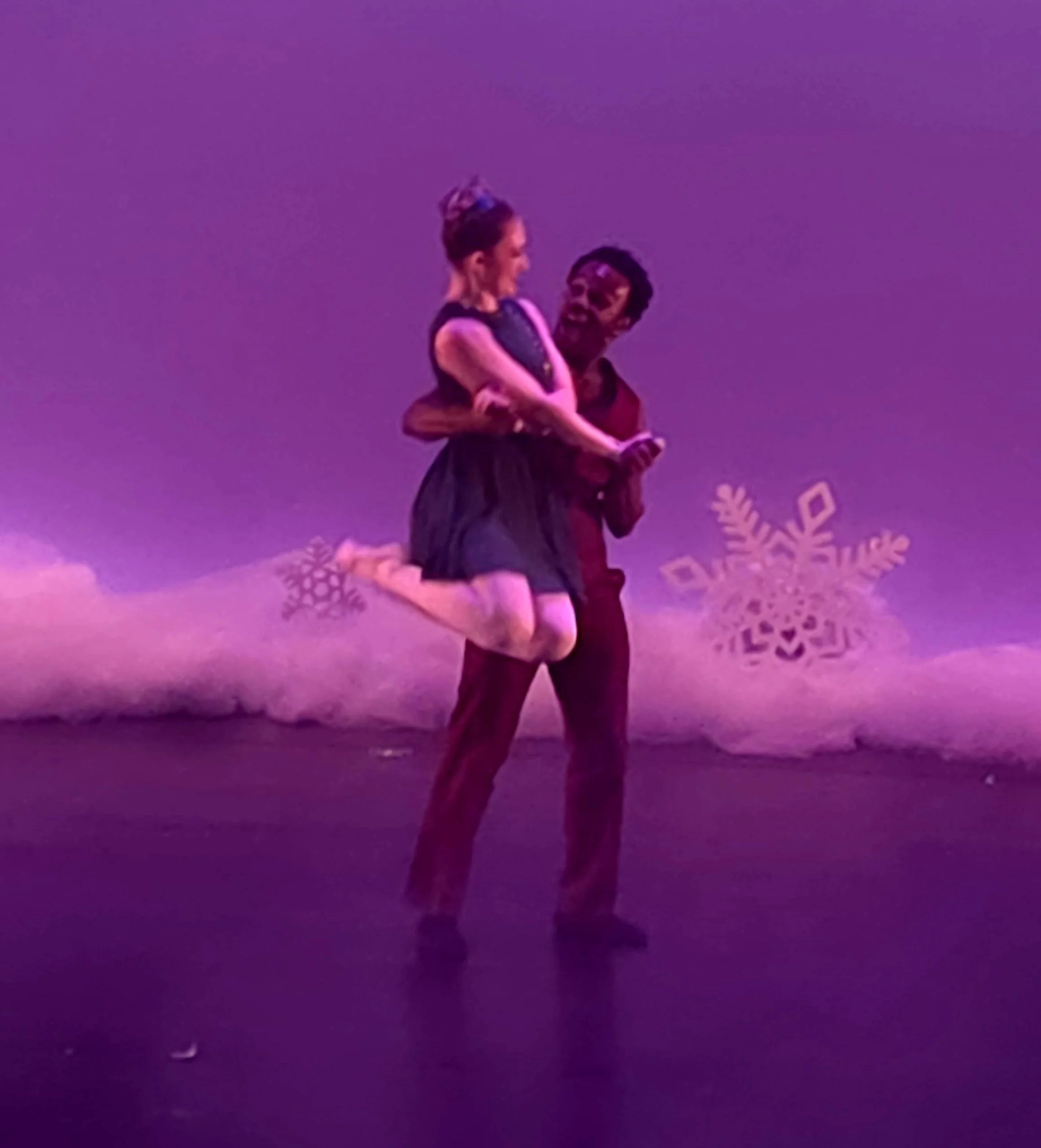 Two ballet dancers performing on stage with a purple background and snowflake decorations.