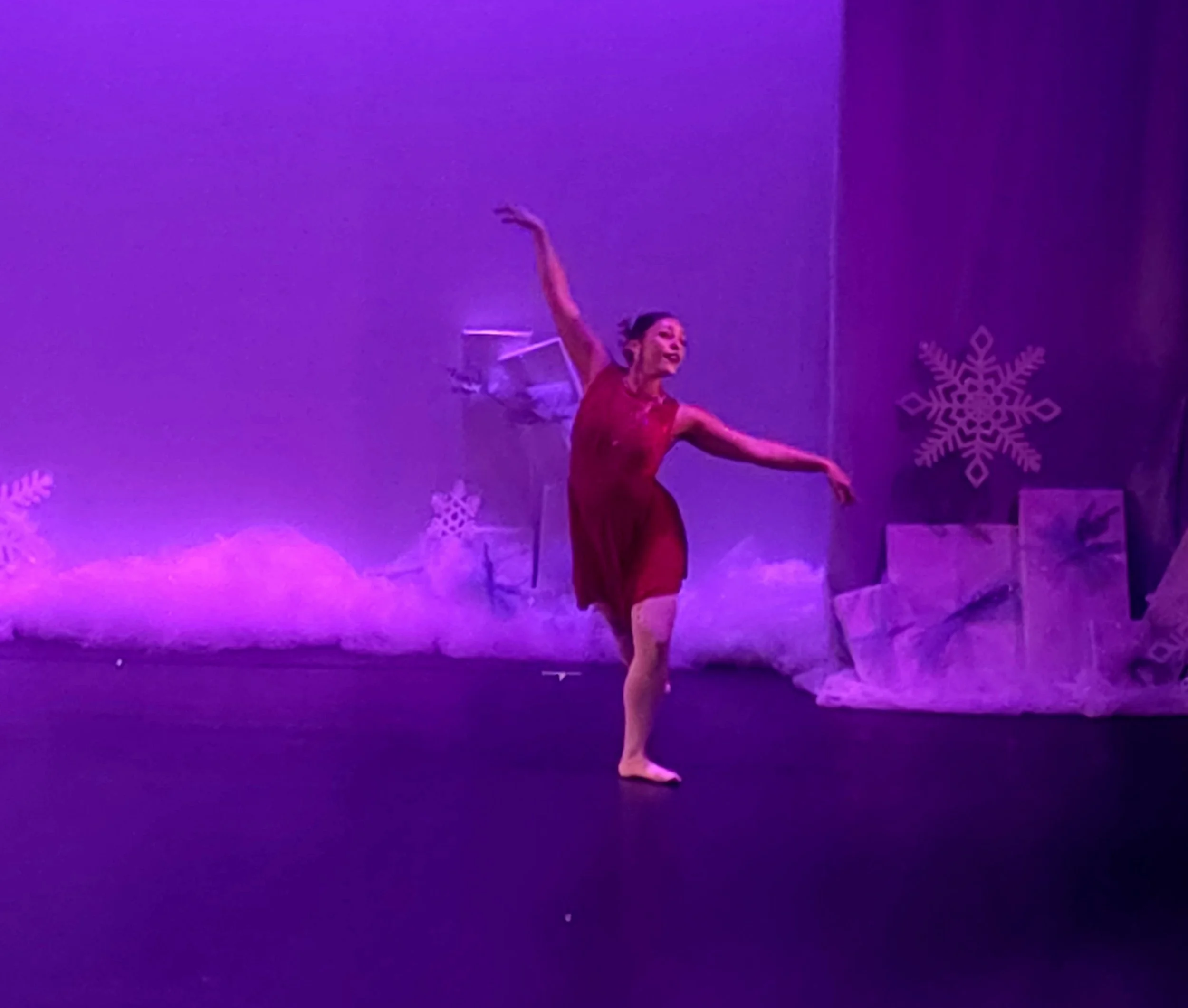 A female ballet dancer performing on stage under purple lighting, wearing a red dress with snowflake decorations in the background.