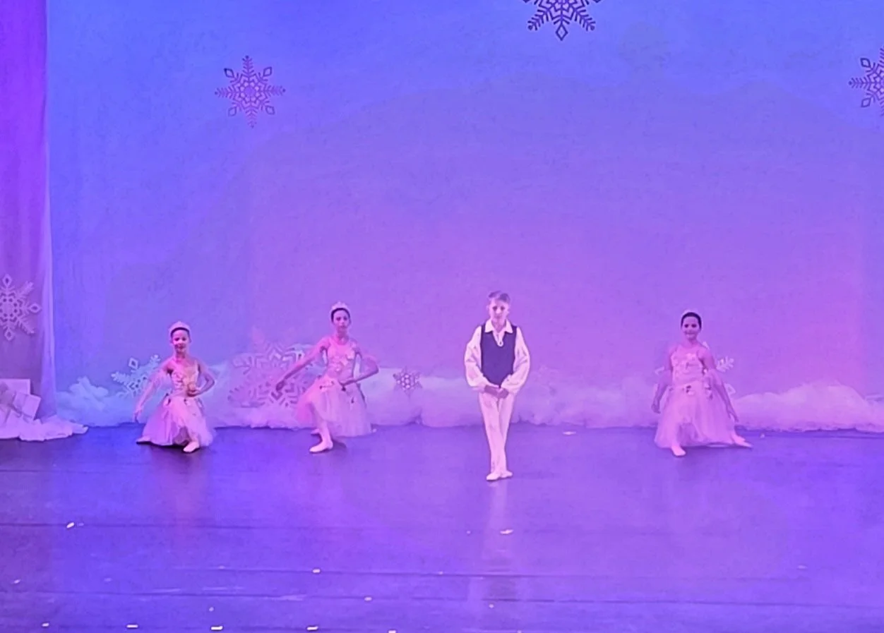 Young children performing a ballet on stage with a winter snowflake-themed backdrop and purple lighting.