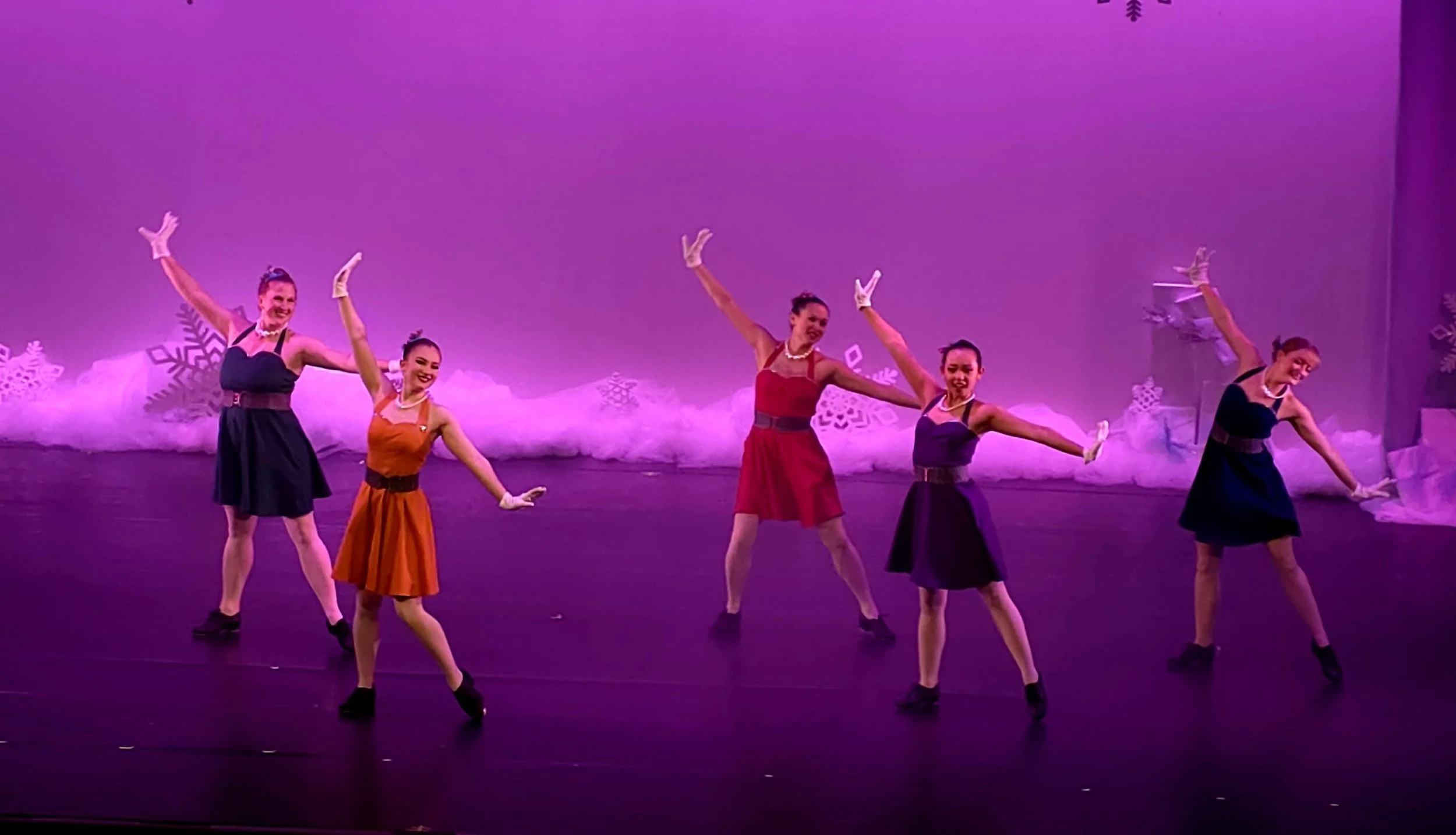 Five women in vintage-style dresses performing a dance on stage with a purple backdrop and snowflake decorations.