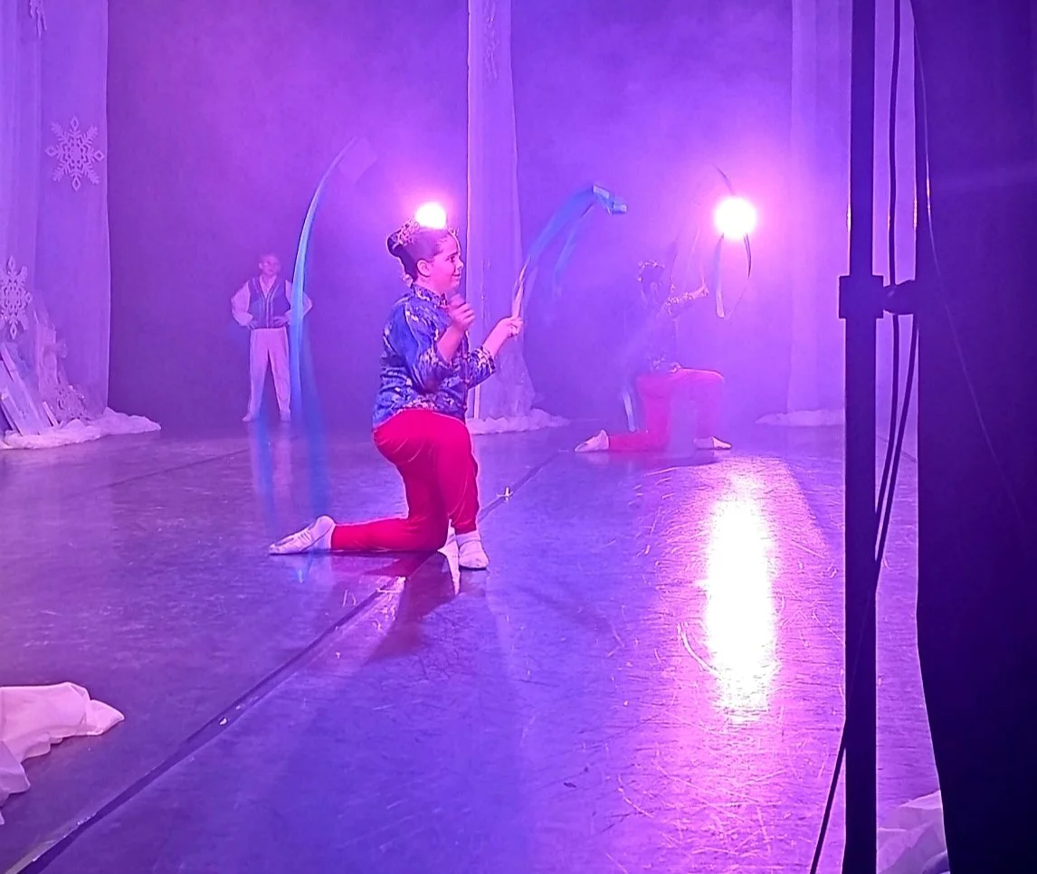 Child performing a dance on stage surrounded by purple lighting, with other children in costumes in the background.