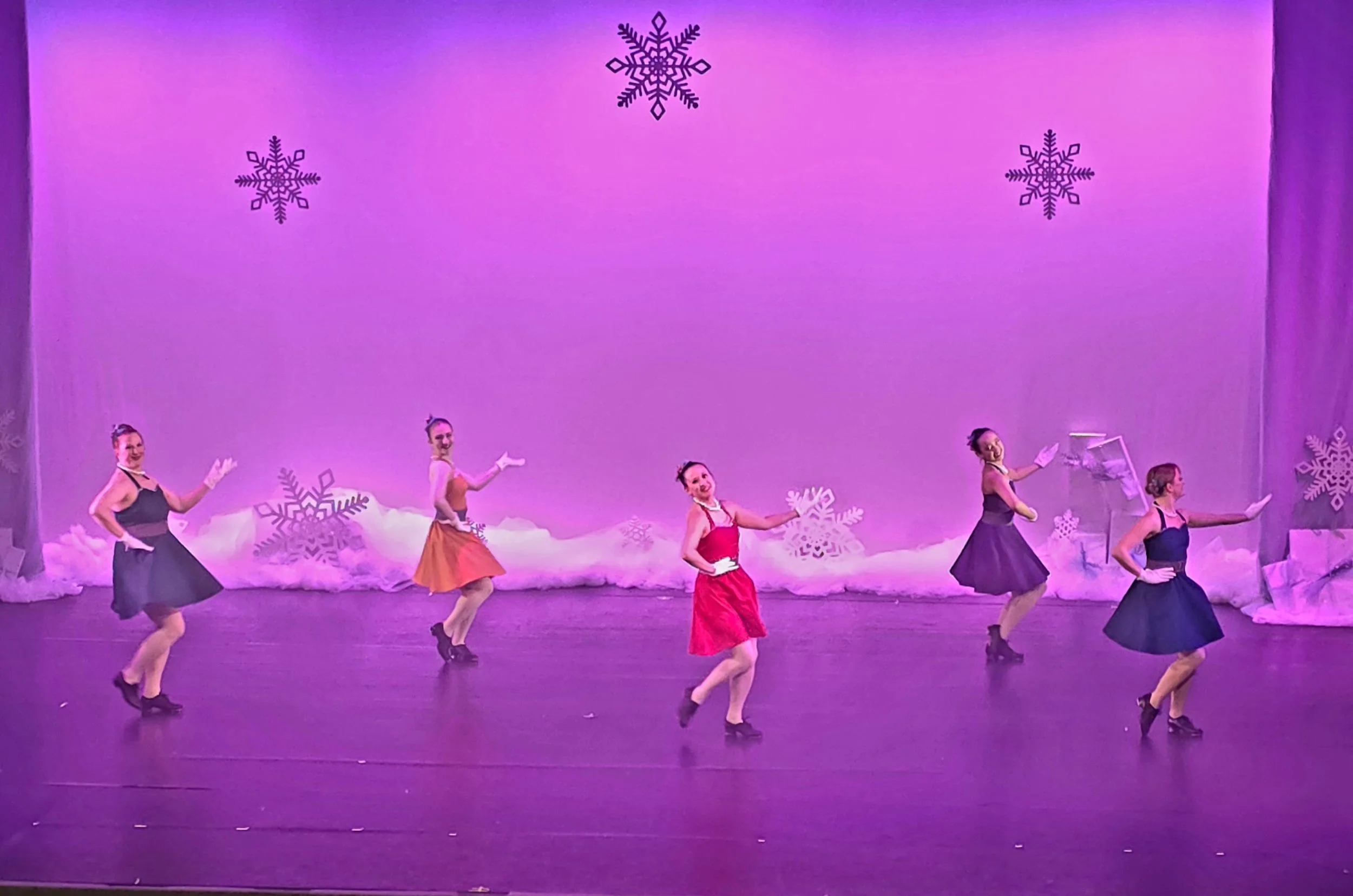 Ballet dancers in colorful dresses perform on stage with a purple background decorated with snowflakes and clouds.