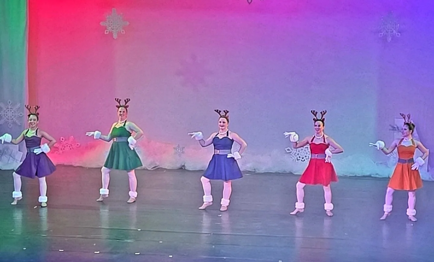 Six female dancers in colorful dresses with reindeer antler headbands and white gloves, performing on stage with snowflake decorations in the background.