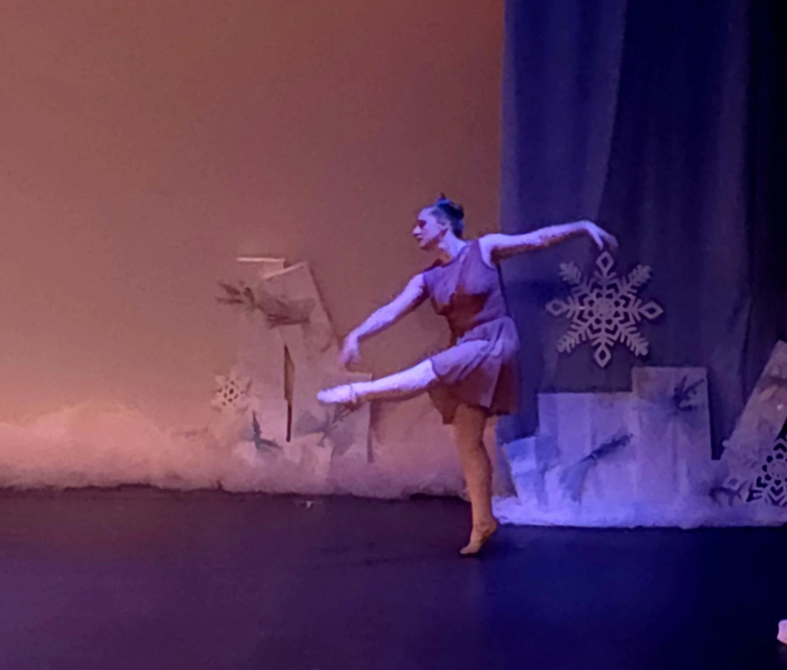 A dancer performing on stage with lighting creating a colorful, smoky atmosphere. The dancer is dressed in a costume, striking a pose, with a large snowflake ornament on the floor nearby.