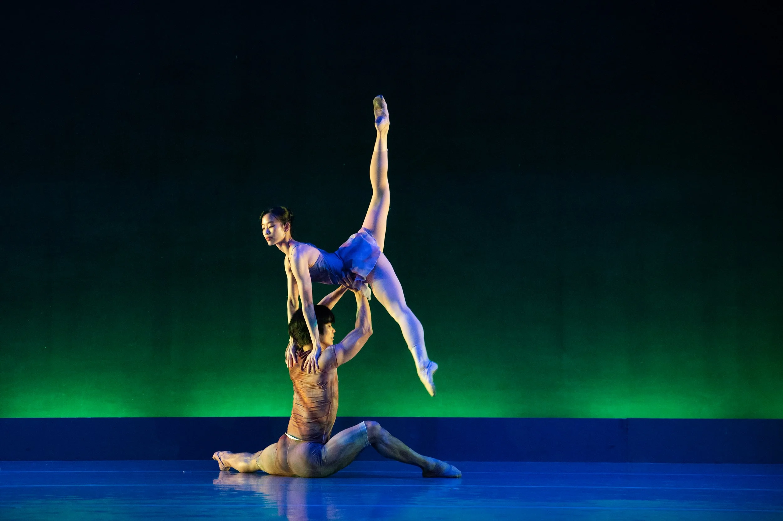 Two ballet dancers perform an acrobatic pose on stage with a dark background and green lighting. One dancer is in a handstand on the other's shoulders, while the second dancer is in a pose with one leg extended upward and the other bent, supporting t
