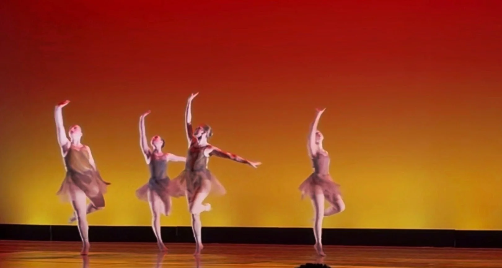 Five ballet dancers performing on stage with a yellow-orange background, each in a different pose with arms raised and wearing brown tutus.