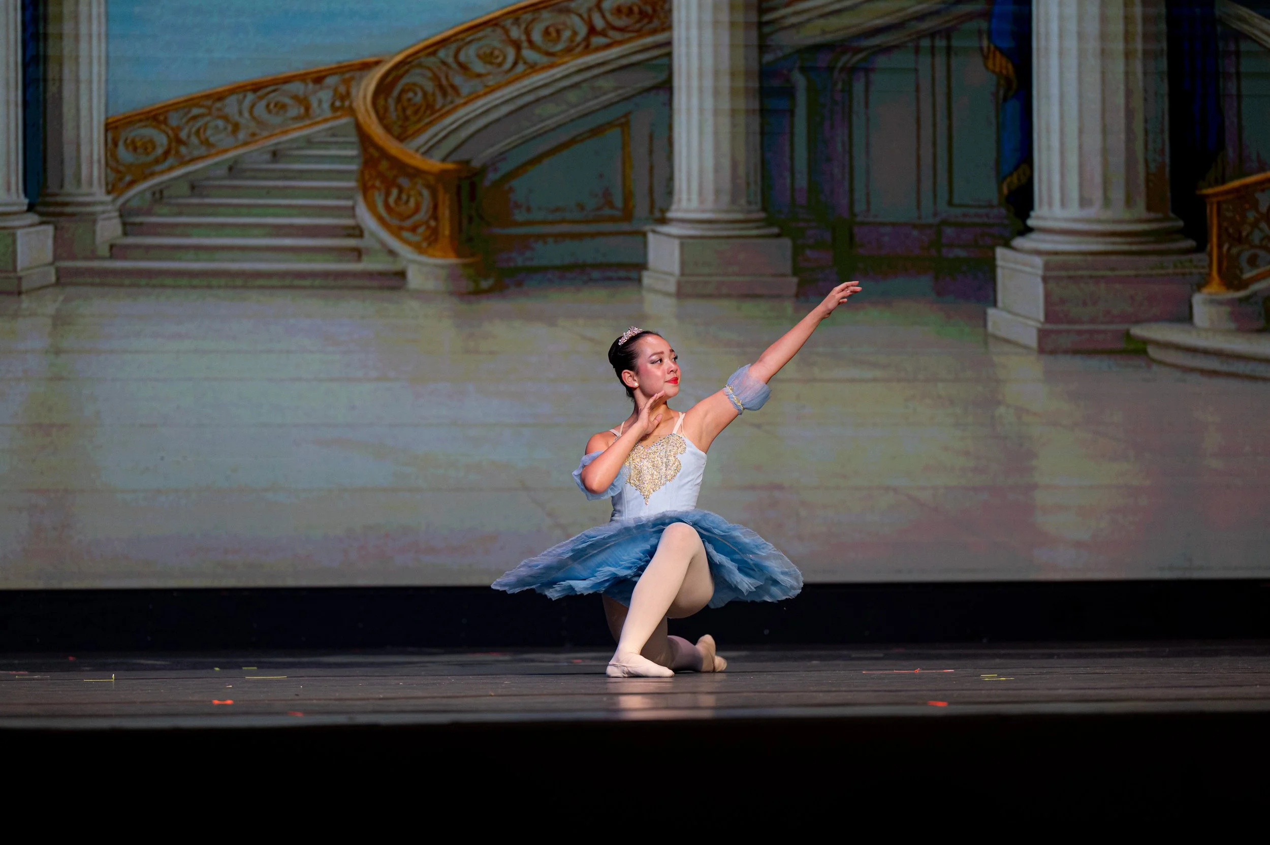 A ballerina in a blue tutu performs a dance on stage, with a grand staircase and ornate decor painted as a backdrop.