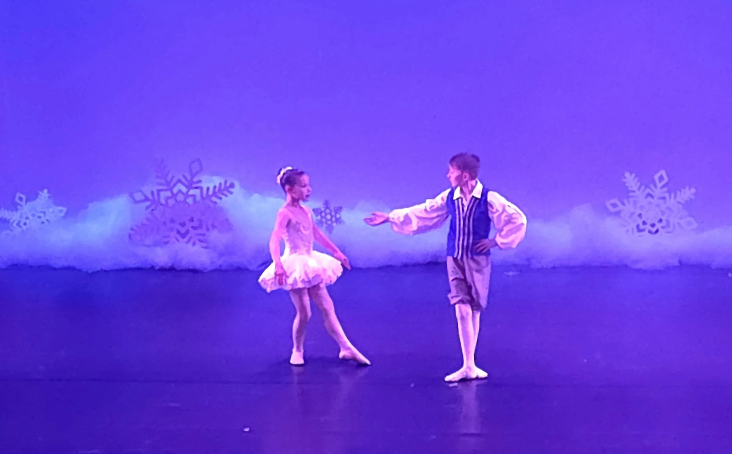 Two children, a girl and a boy, perform a ballet on stage with snowflake decorations in the background, under purple lighting.