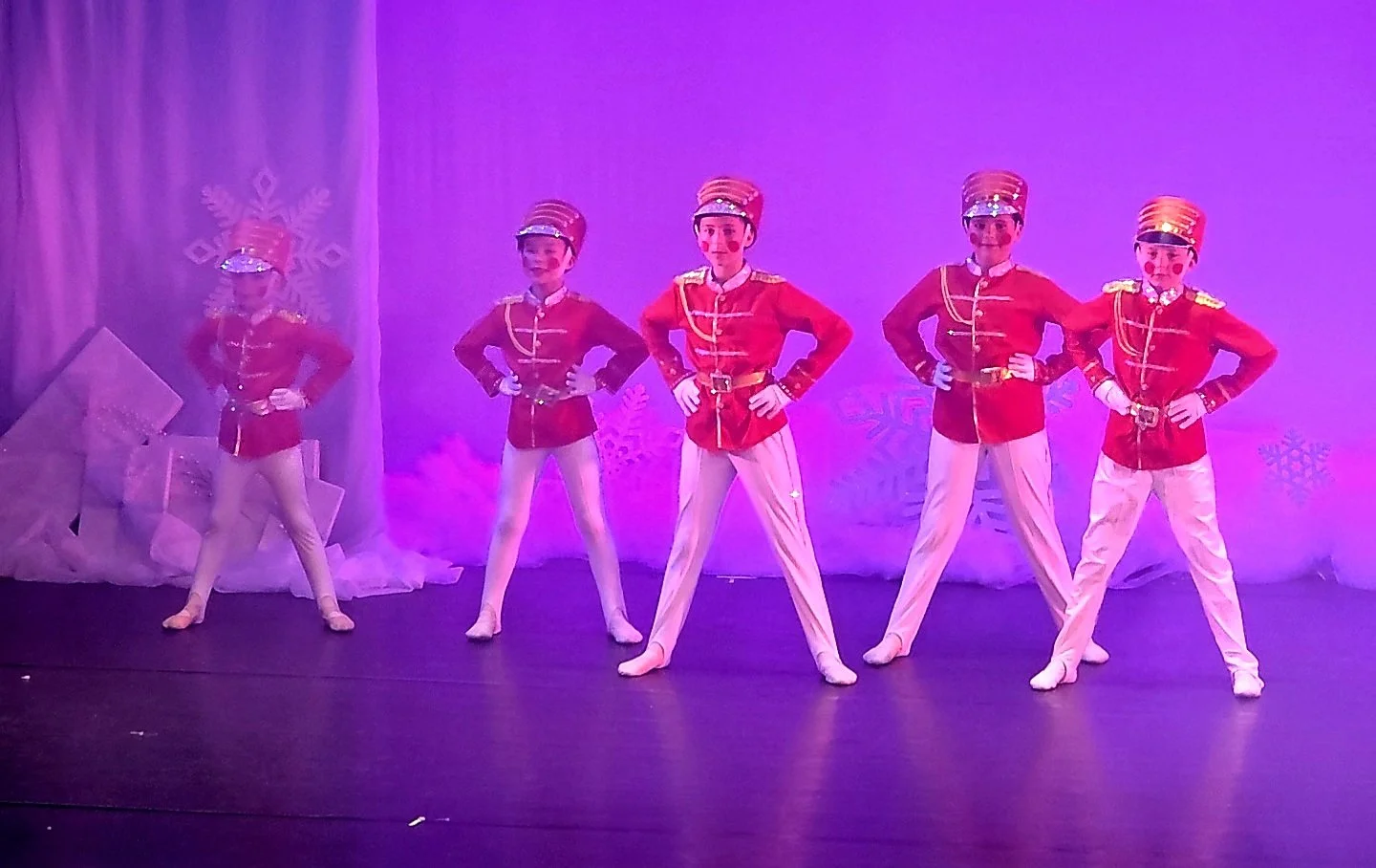 Five children dressed as toy soldiers perform a dance on stage with a festive winter background.