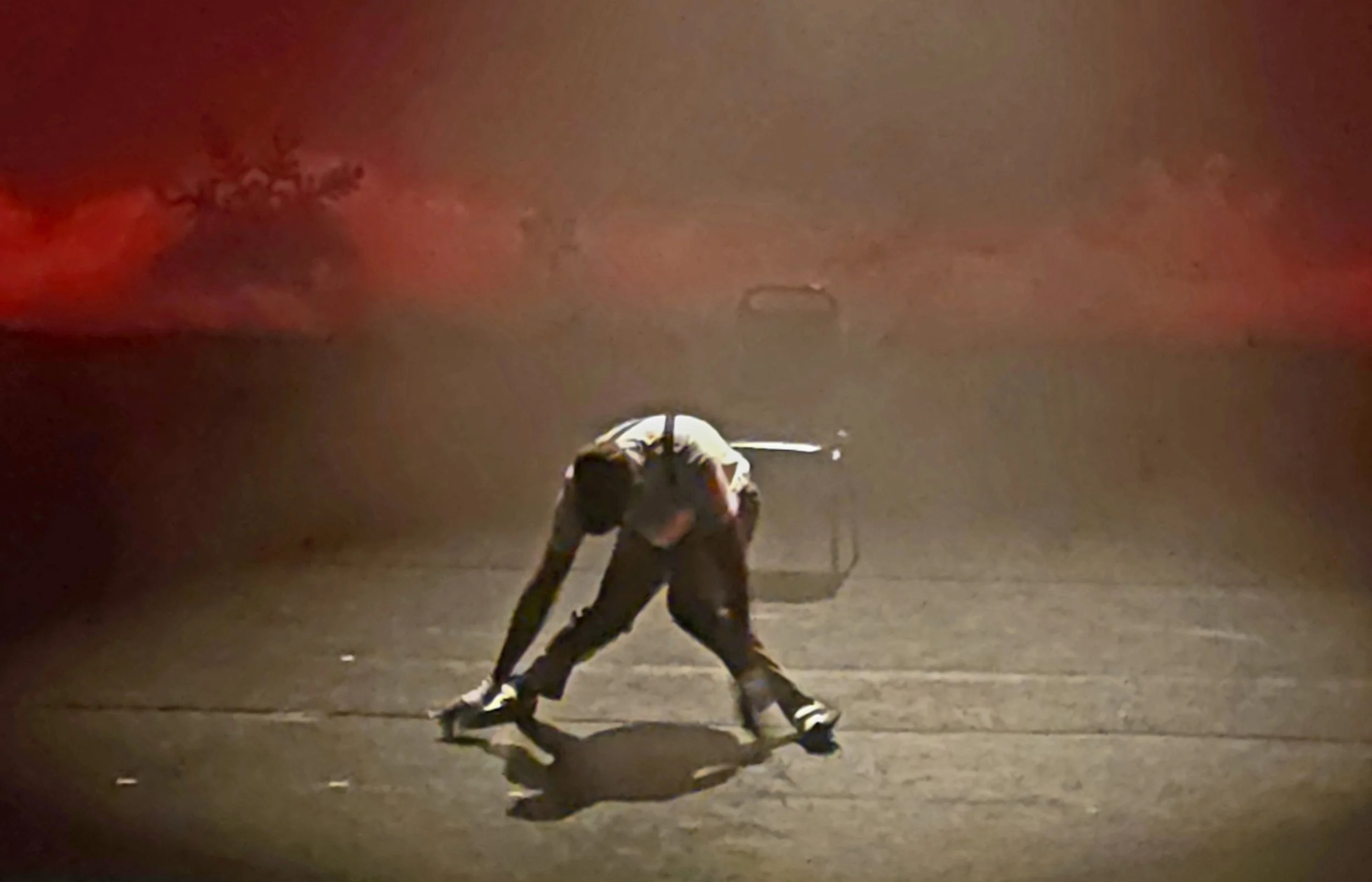 A person crouching and leaning forward on a dark street at night, with a faint red glow in the background.