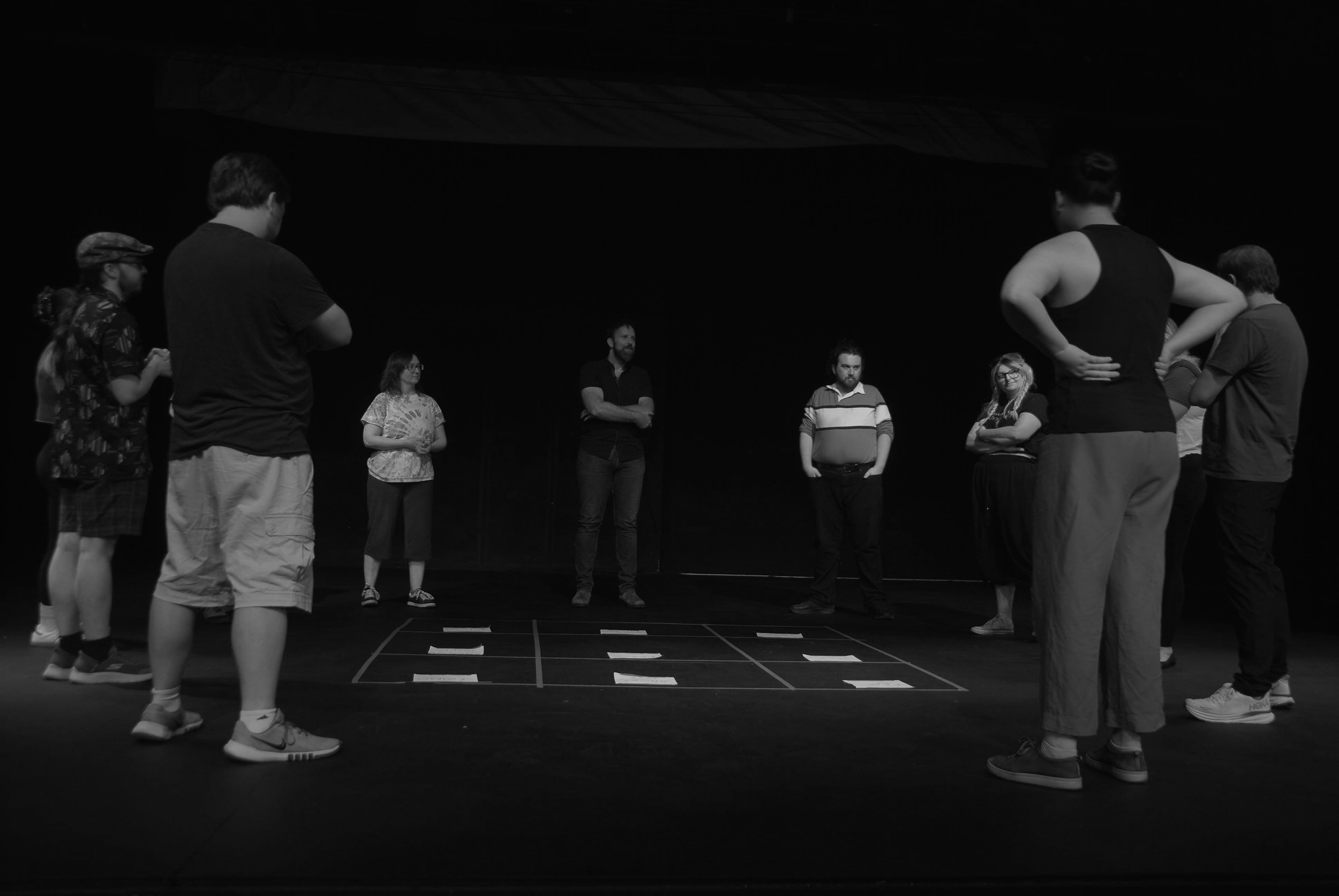 A black-and-white photograph of a diverse group of actors in casual rehearsal clothes standing on a stage. The floor is marked with white tape 'spikes' to indicate blocking. The actors are positioned in a semi-circle.