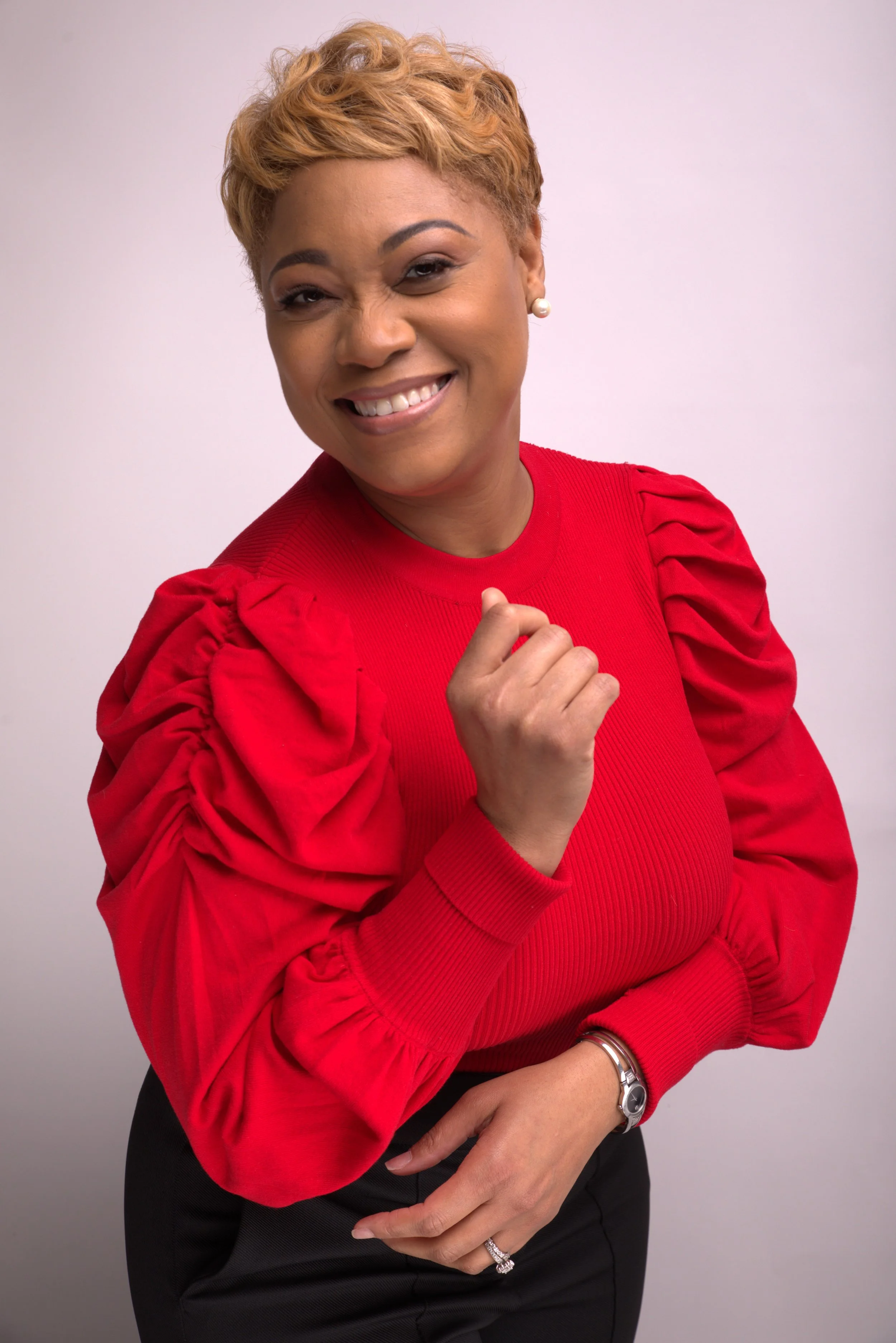 A woman with short blonde hair smiling and wearing a red blouse with puffed sleeves and black pants, standing against a plain background.