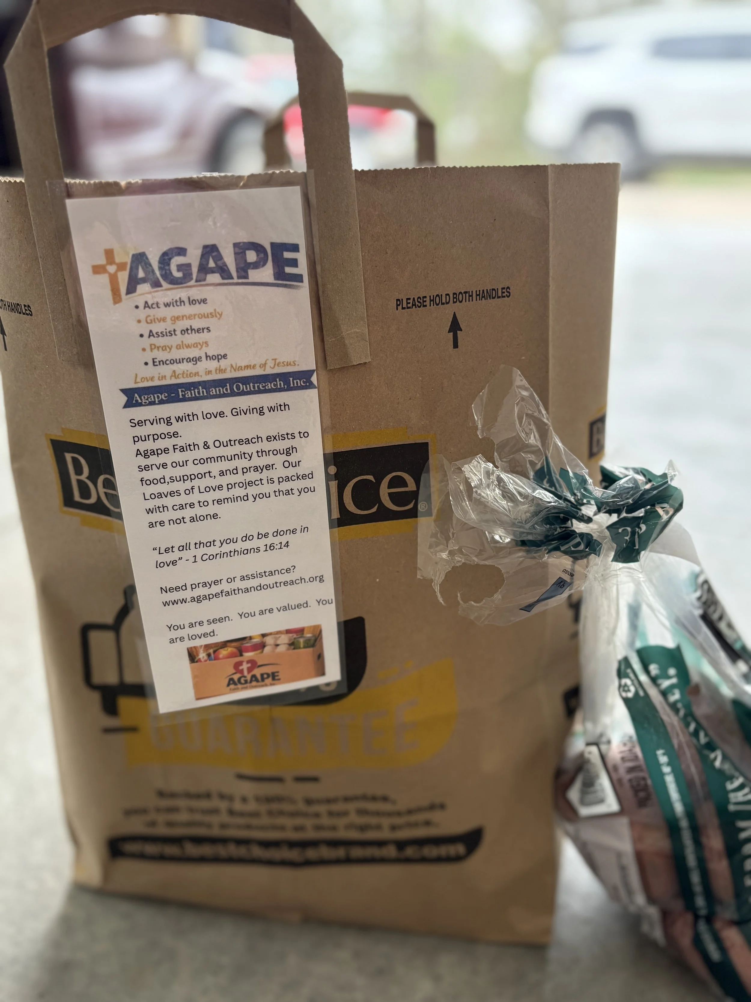 A brown paper bag with a label from Agape Faith and Outreach Inc., containing food, and a crumpled white and green plastic bag nearby. The background shows a parking lot with cars.