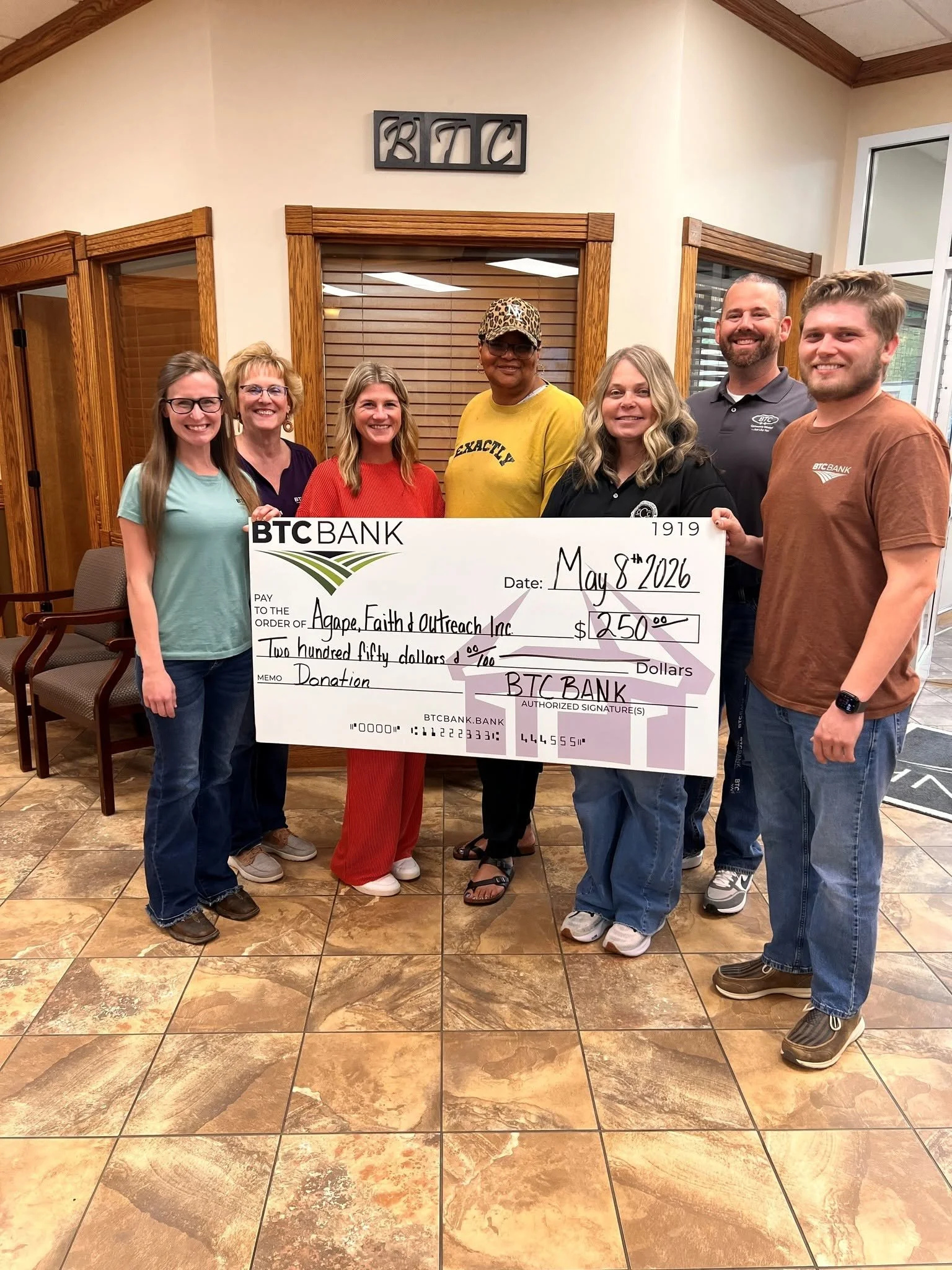 Group of seven people standing indoors holding a large check made out to Agape Faith Outreach Inc. for $250, dated May 8, 2026, with the BTC Bank logo. They are smiling, and the group includes four women and three men. The background features wooden 