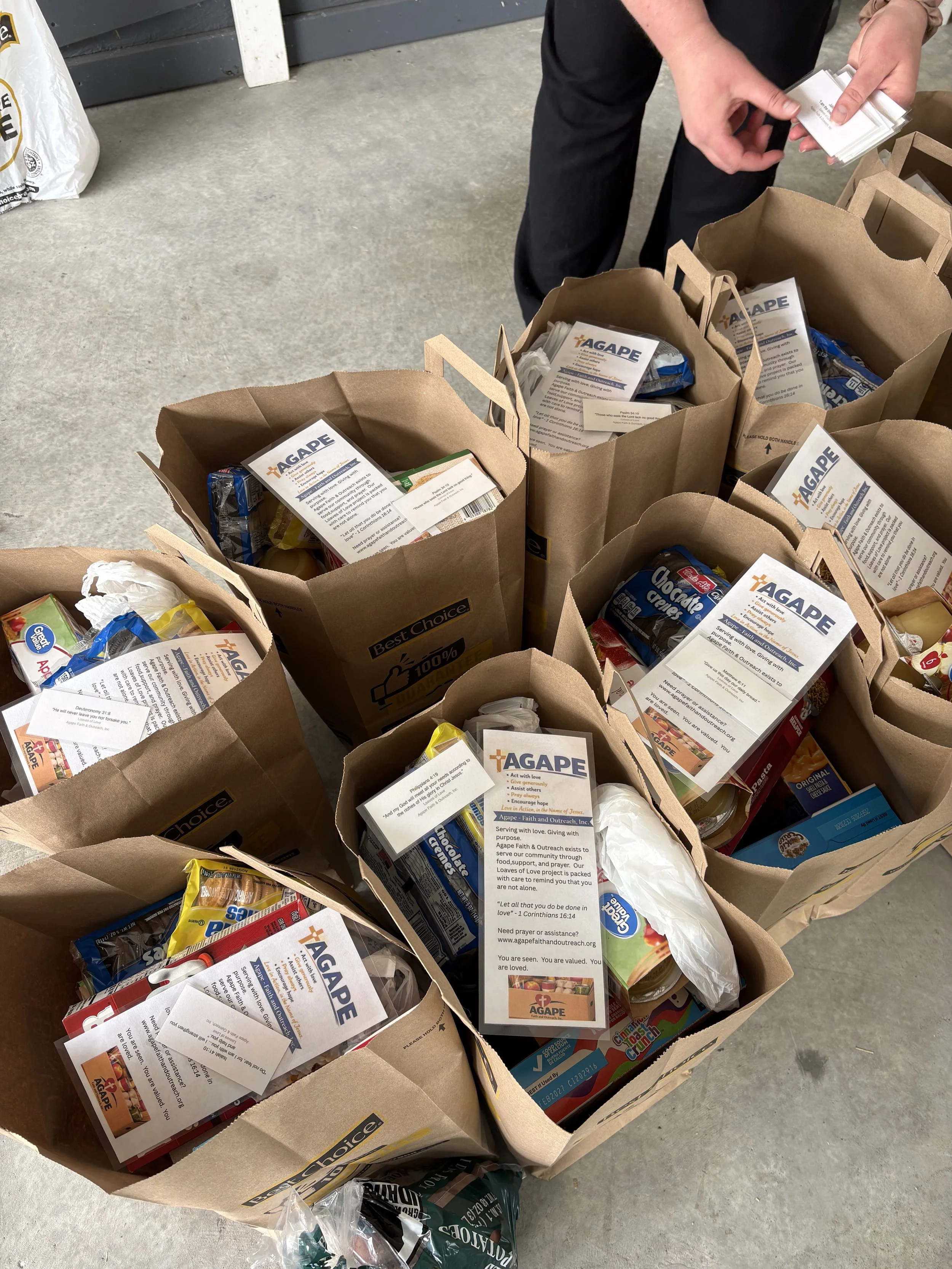 Multiple brown paper grocery bags filled with food items and informational flyers from AGAPE, a charity organization, on a concrete floor. A person's hands are seen holding a pamphlet, and various food packages are visible inside the bags.