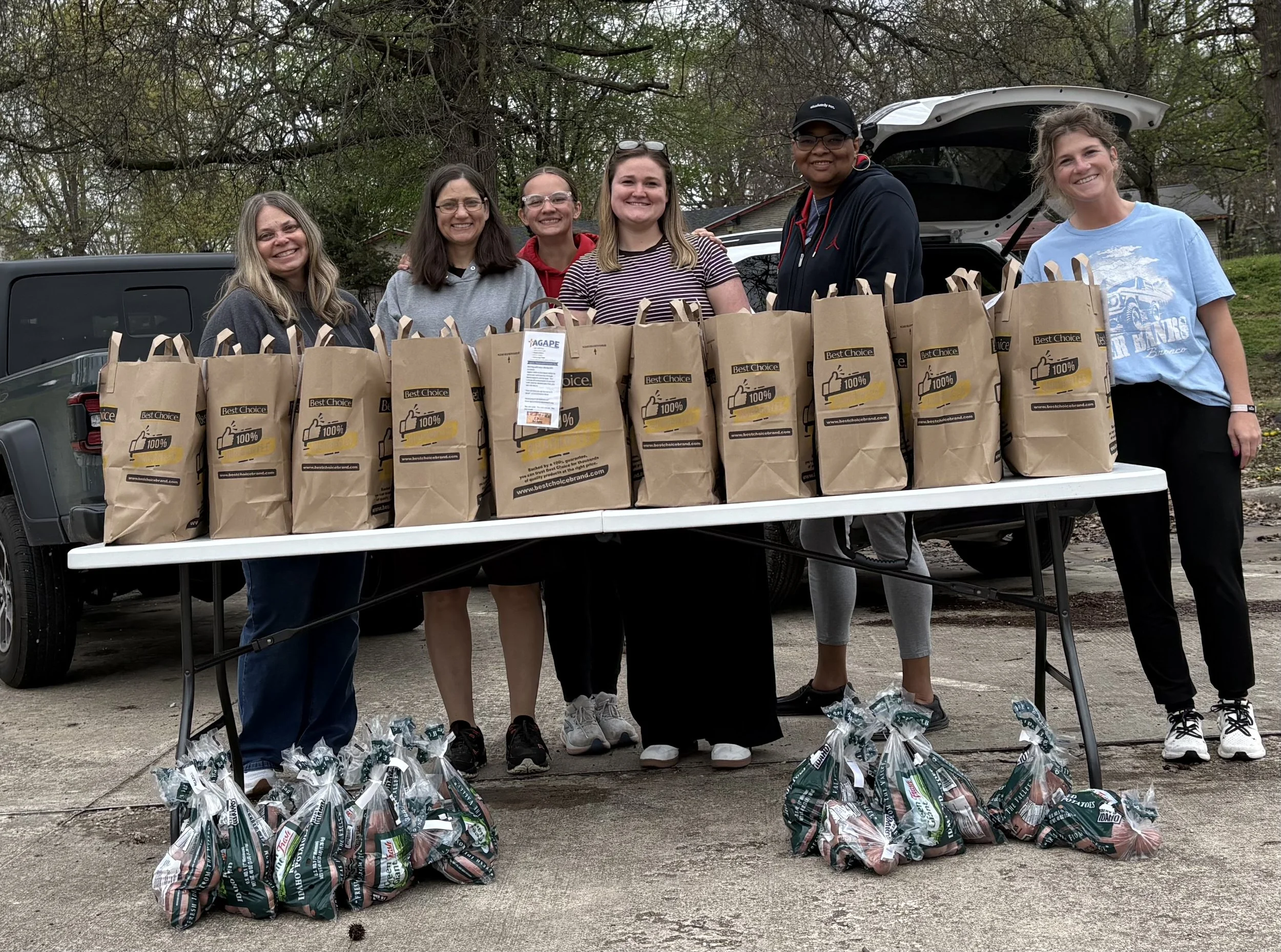 Group of people standing behind a table filled with grocery bags, with more bags on the ground, outdoors with trees in the background.