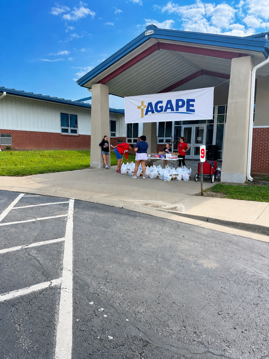 People gathering food donations under a large banner with the word 'AGAPE' outside a building on a sunny day.