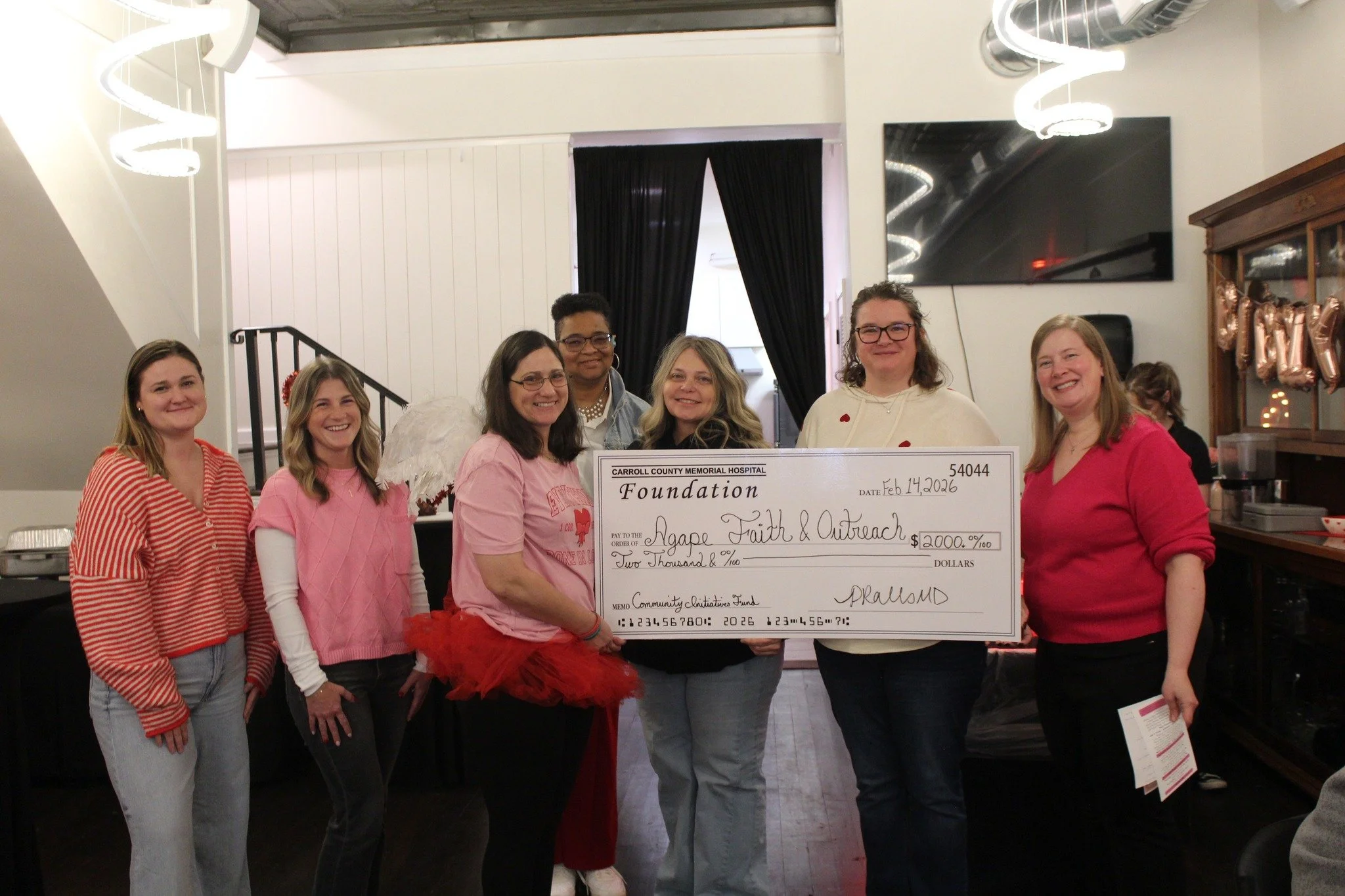 Group of seven women standing together indoors, holding an oversized check for $2000 made out to Agape Faith & Outreach, with decorated balloons in the background for a donation event.