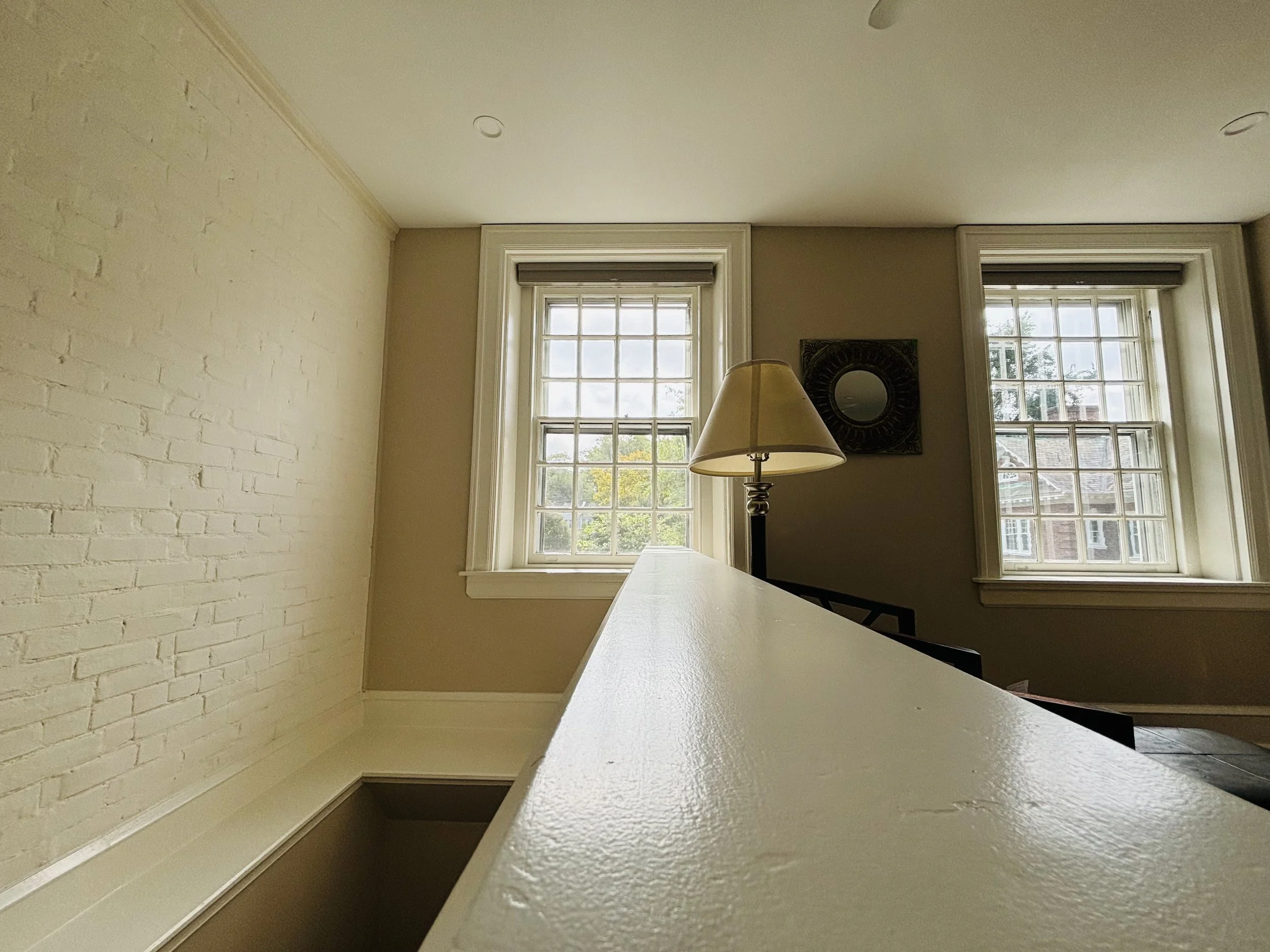 Interior view of a room with two large windows, a wall-mounted mirror, a standing lamp, and a railing or half-wall in the foreground.