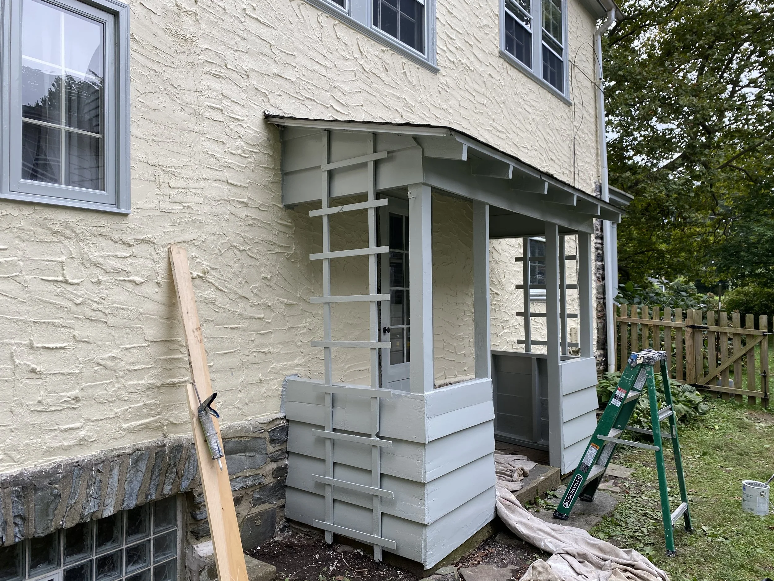Construction of a small porch or entryway with white painted wood, attached to a yellow brick house, with a ladder and construction materials nearby.
