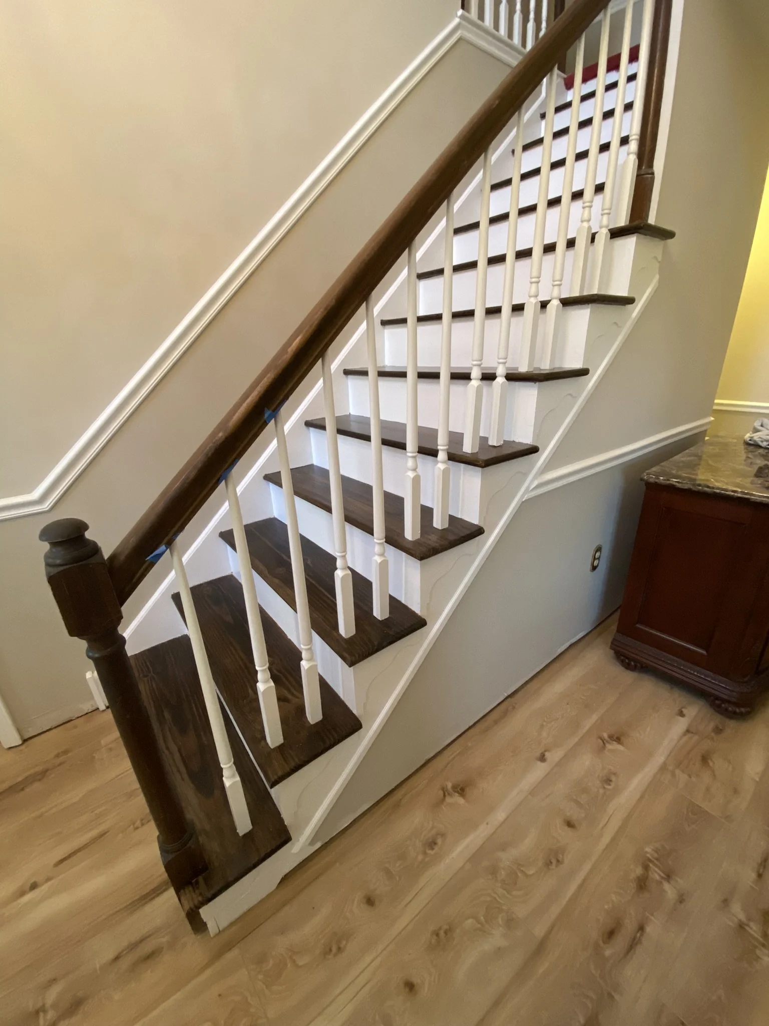 Interior view of a staircase with dark wooden steps, white spindles, and a dark wooden handrail, next to a beige wall and a wooden cabinet with a marble countertop.