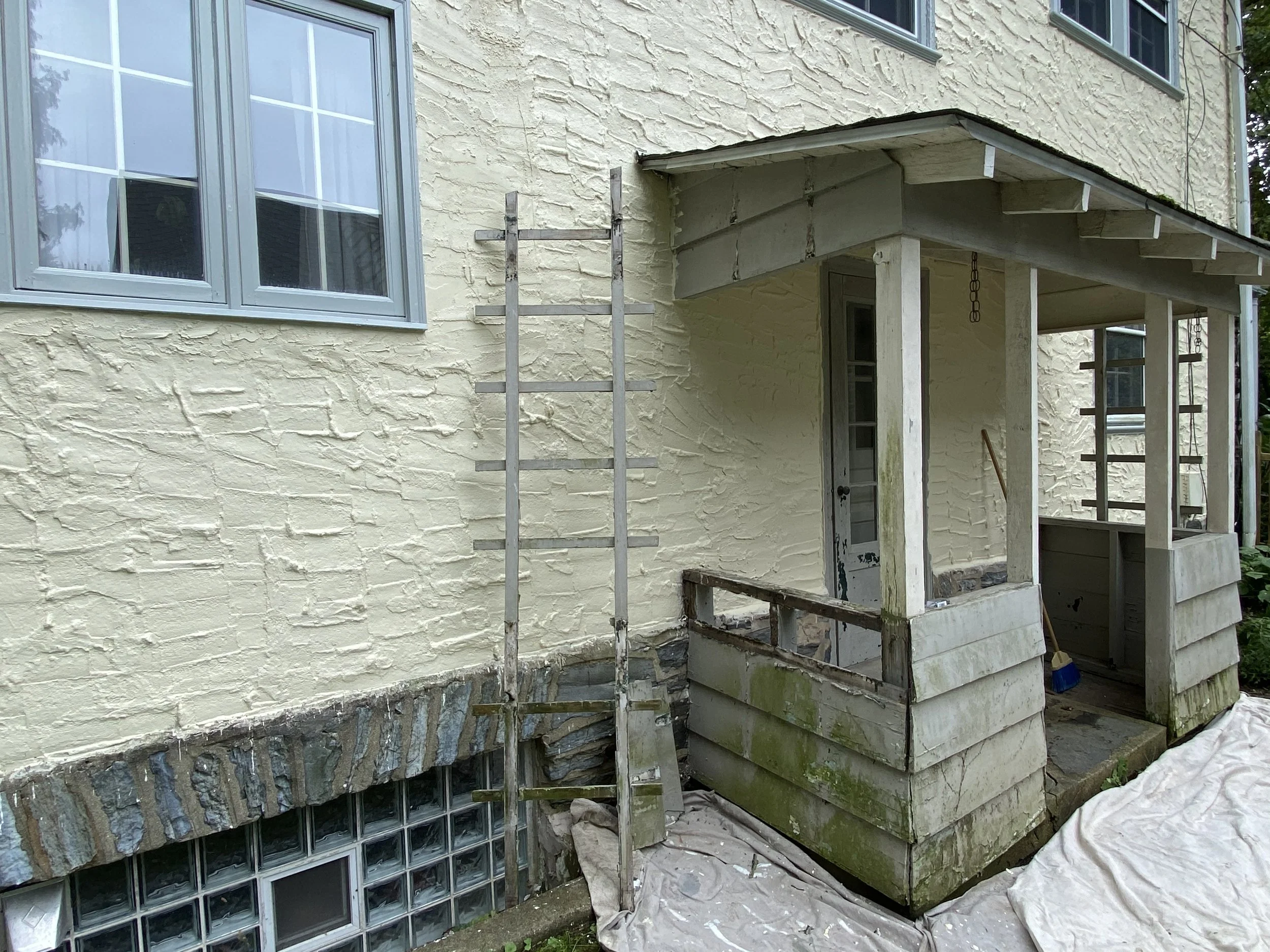 A house exterior with a small porch under renovation, showing scaffolding and weathered wooden railing, sunlit yellow textured wall, and a window with blue trim.