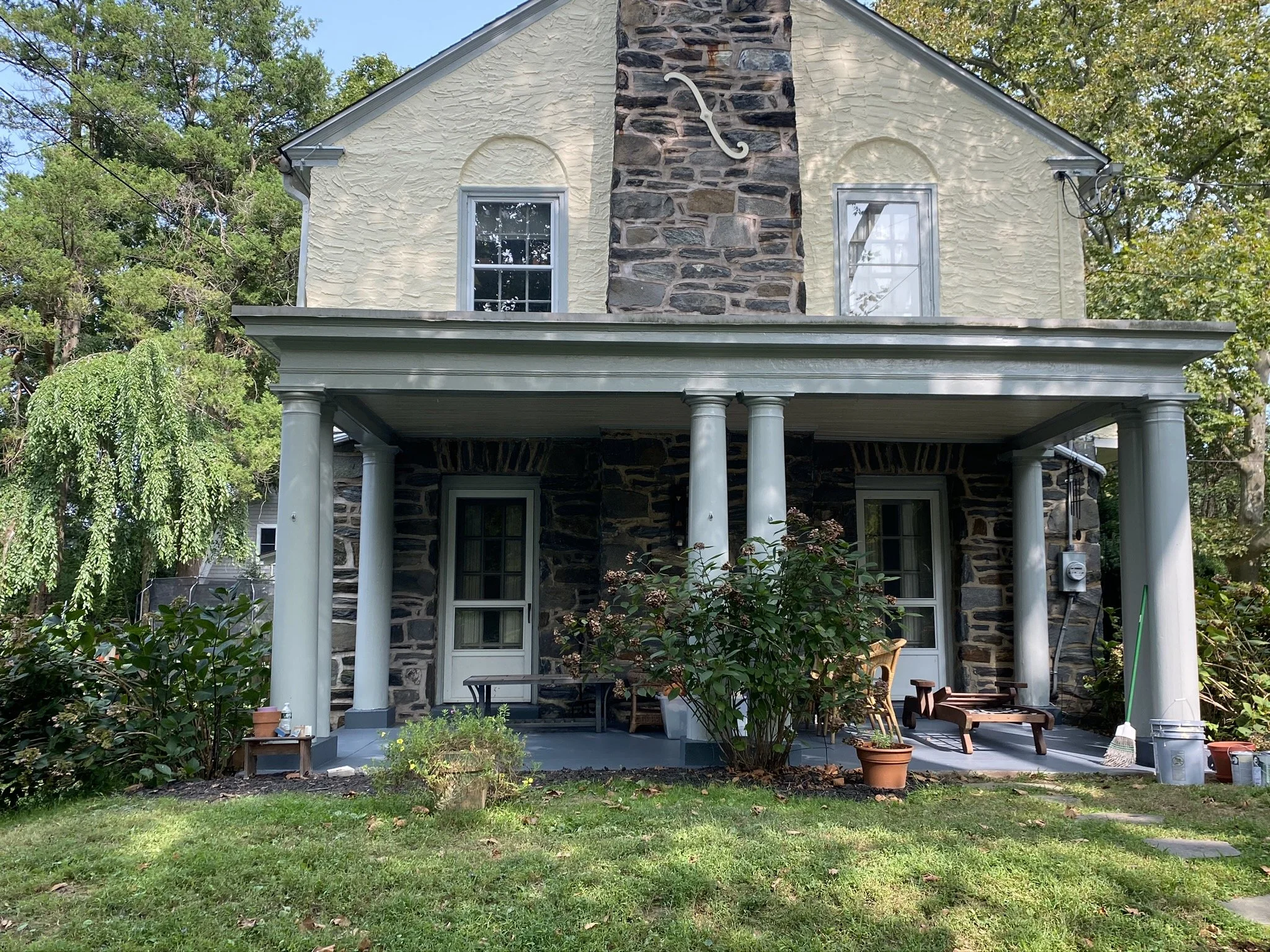 A two-story house with a stone chimney and a porch, featuring two windows on the upper floor, surrounded by trees and greenery.