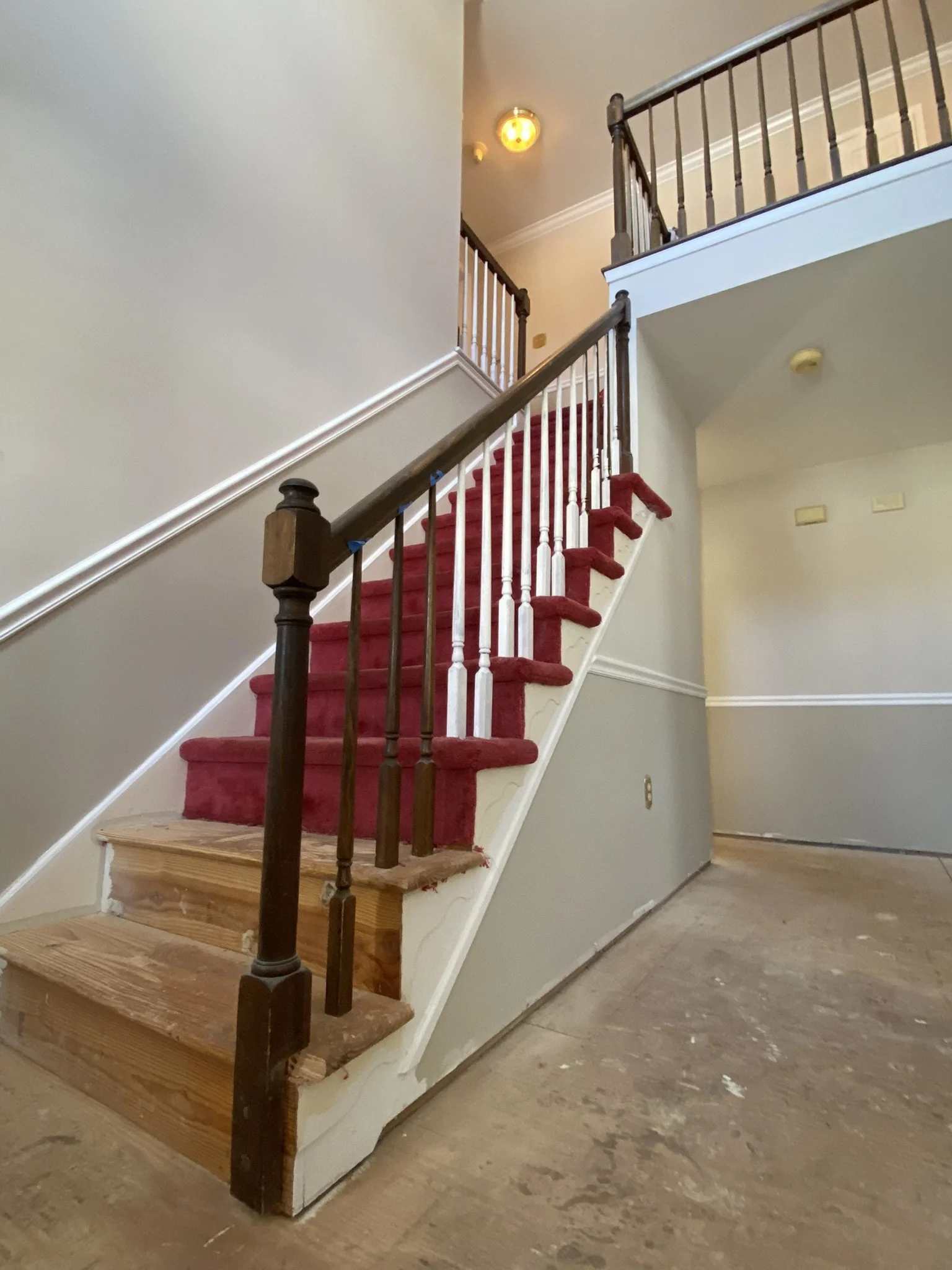 The interior of a house with a staircase containing red carpet runners, wooden steps, and a wooden and white banister, leading to the upper floor. The staircase is partially renovated, with some unfinished wooden steps at the bottom and a beige wall 