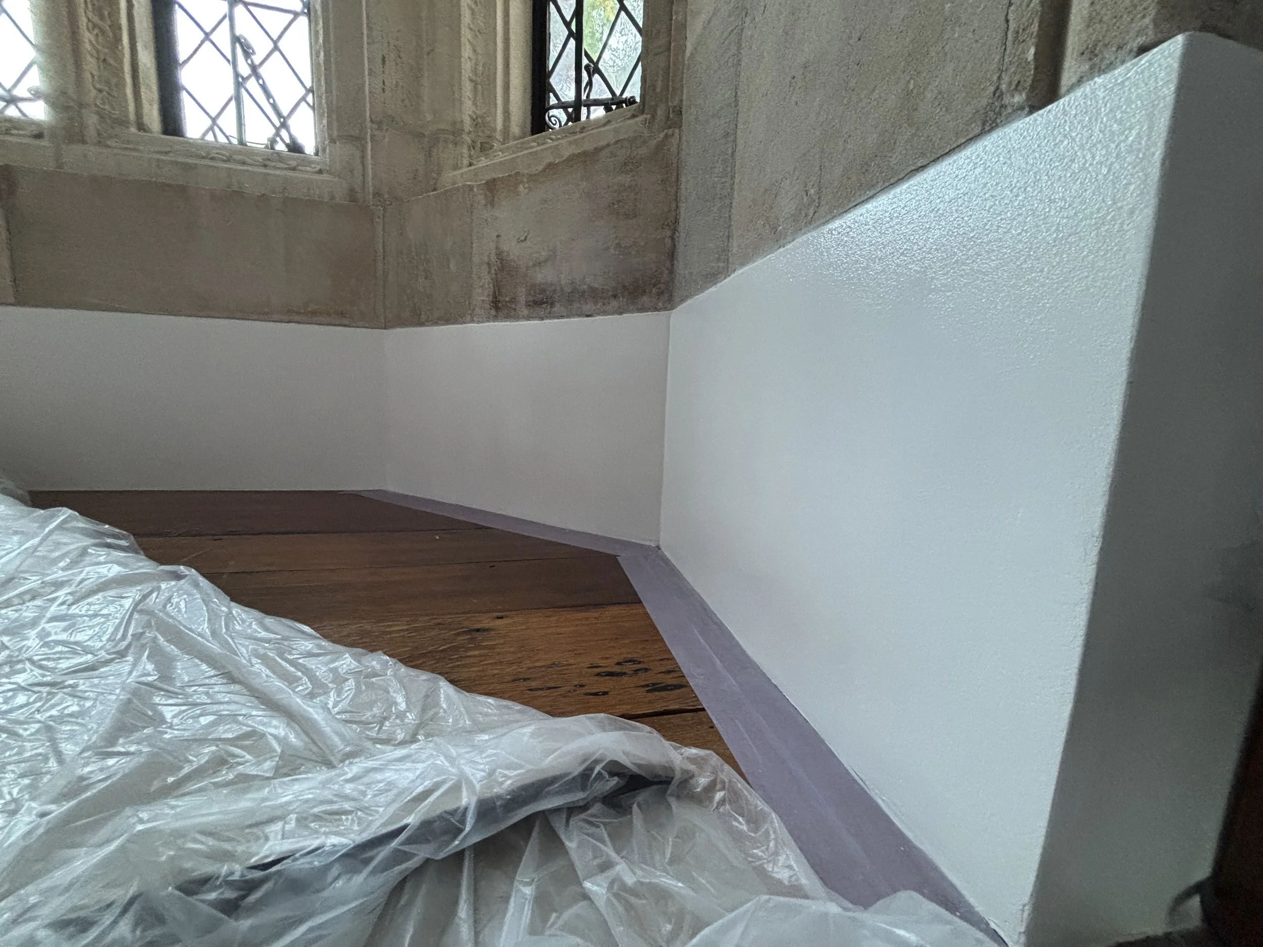 Interior corner of a room with a stone wall and windows, showing a section of a freshly painted white wall and a wooden floor, with painter's plastic and painter's tape along the floor.