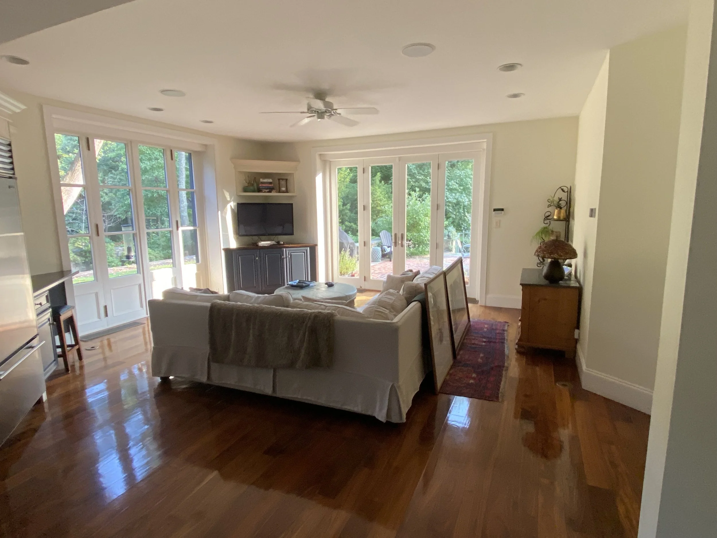 Living room with large windows and glass doors, white sofa, wooden floor, TV, small side tables, decorative items, and outdoor view.