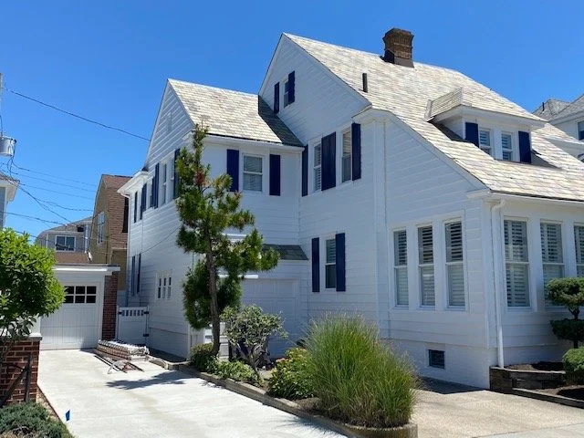 A large white multi-story house with multiple windows and a steep roof, surrounded by a driveway, trees, and landscaped bushes under a clear blue sky.