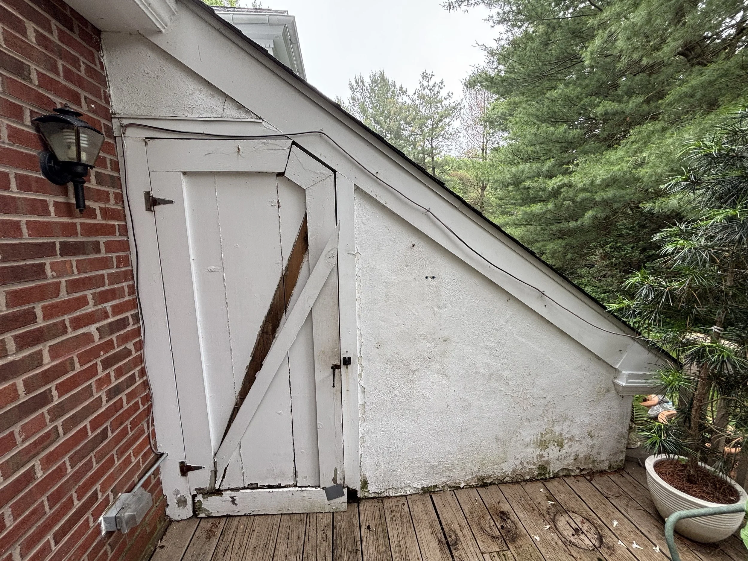 A small wooden shed with a slanted roof, weathered white paint, and a boarded-up door on a wooden deck next to a brick wall and surrounded by green trees.