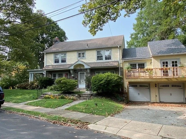 A two-story house with a front porch and greenery, adjacent to a garage with a balcony, on a suburban street with trees and power lines.