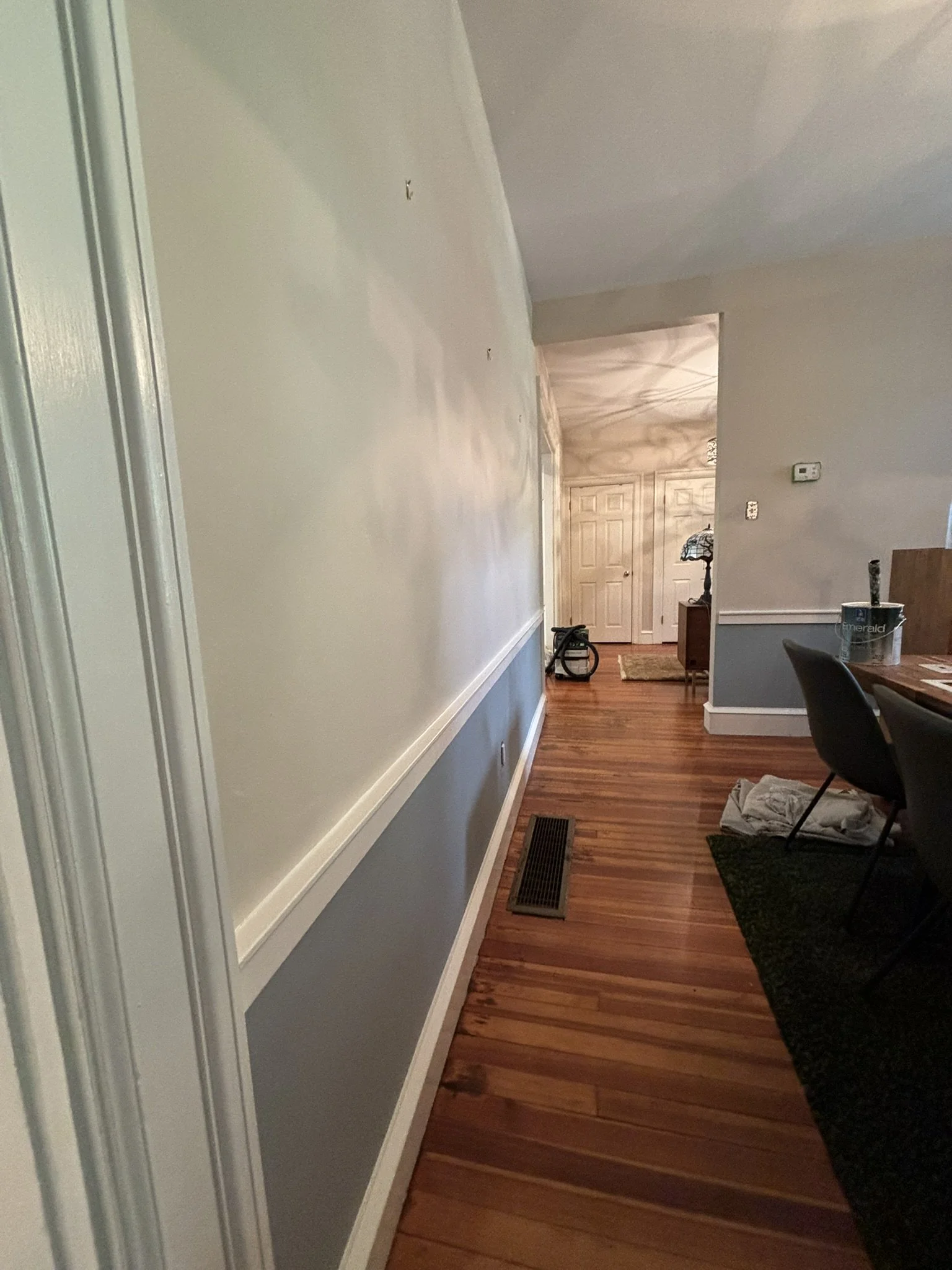 Interior view of a house hallway with hardwood flooring, a vent on the floor, and a partial view of a dining area with chairs. The hallway leads towards a room with closed double doors, with a vacuum cleaner visible near the entrance.