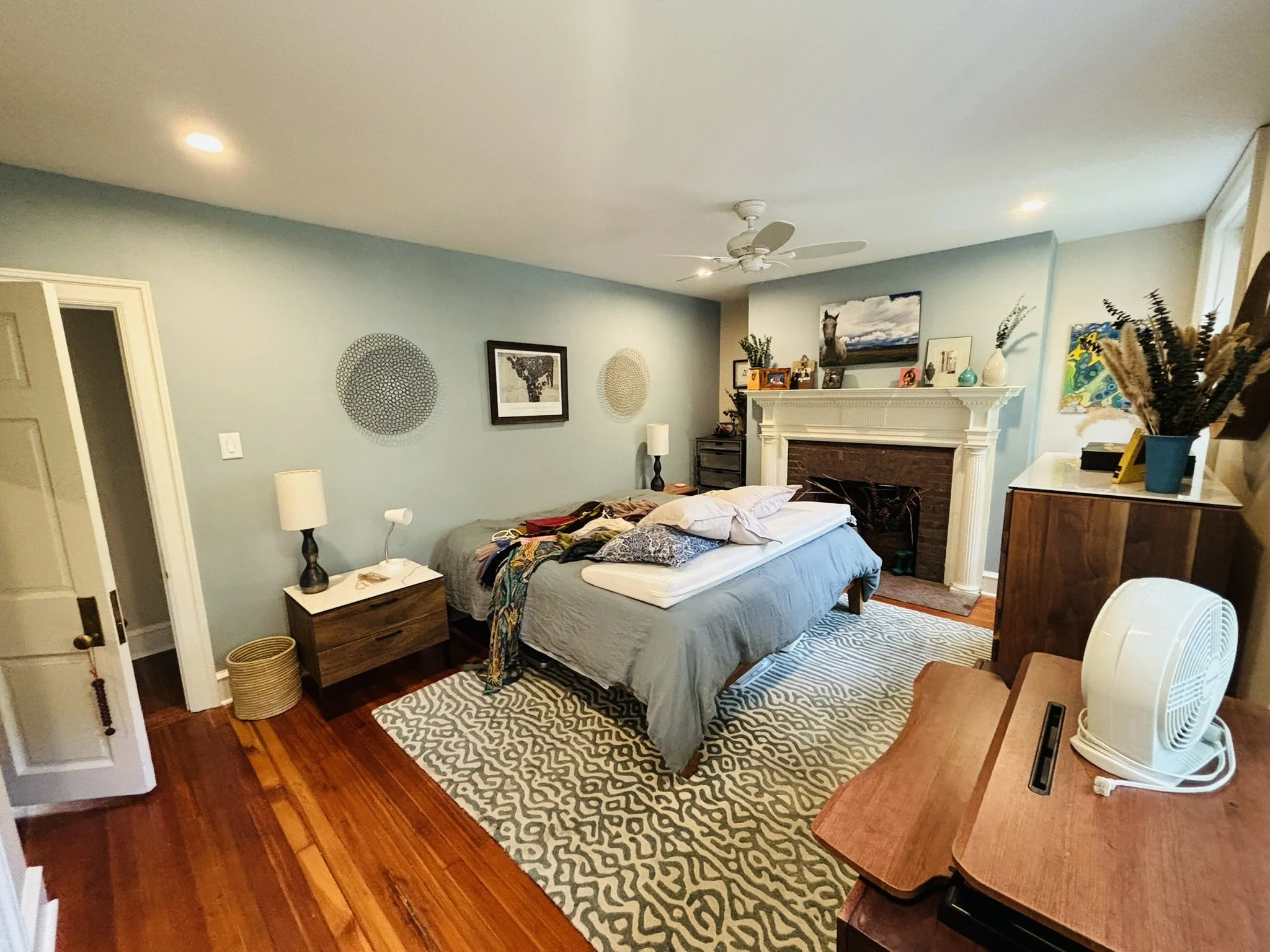 A cluttered bedroom with a bed, nightstand, fireplace, and various decorations including artwork, a clock, and a fan.