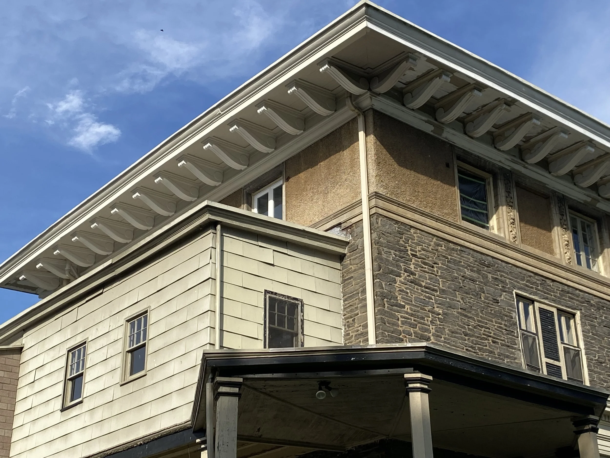 Close-up view of the upper floors of an older house with beige and stone exterior walls, multiple windows, decorative trim, and a roofline against a blue sky with some clouds.