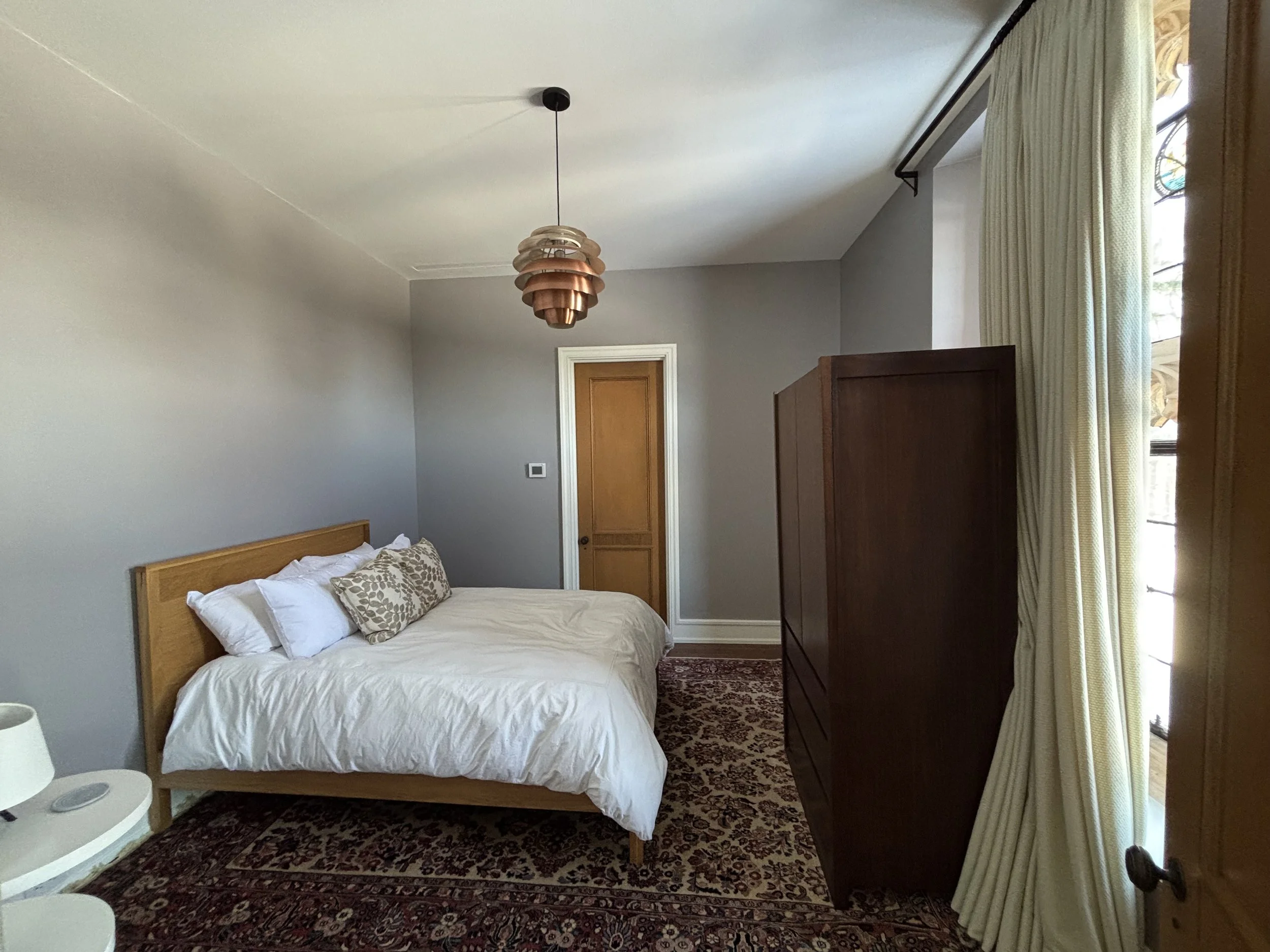 A bedroom with a bed, white bedding, and patterned pillows, a wooden wardrobe, a rug, closed curtains, and a pendant light fixture.