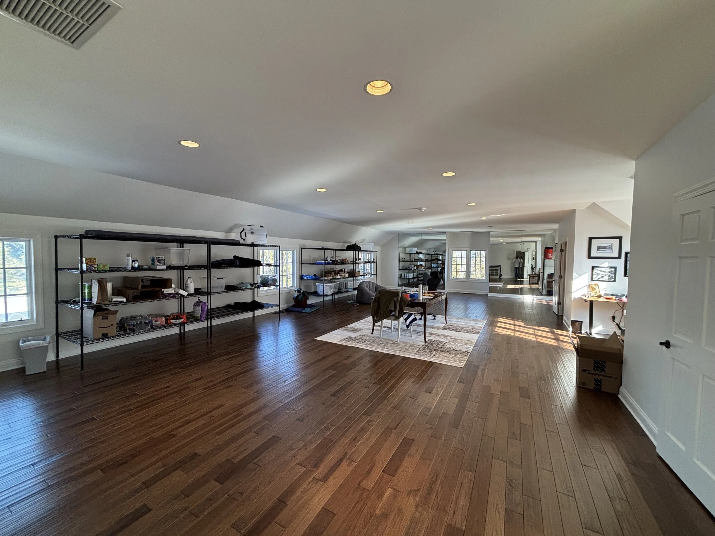 Spacious room with hardwood floors and white walls, containing metal shelving units, a table with chairs, and miscellaneous items, with windows allowing natural light.