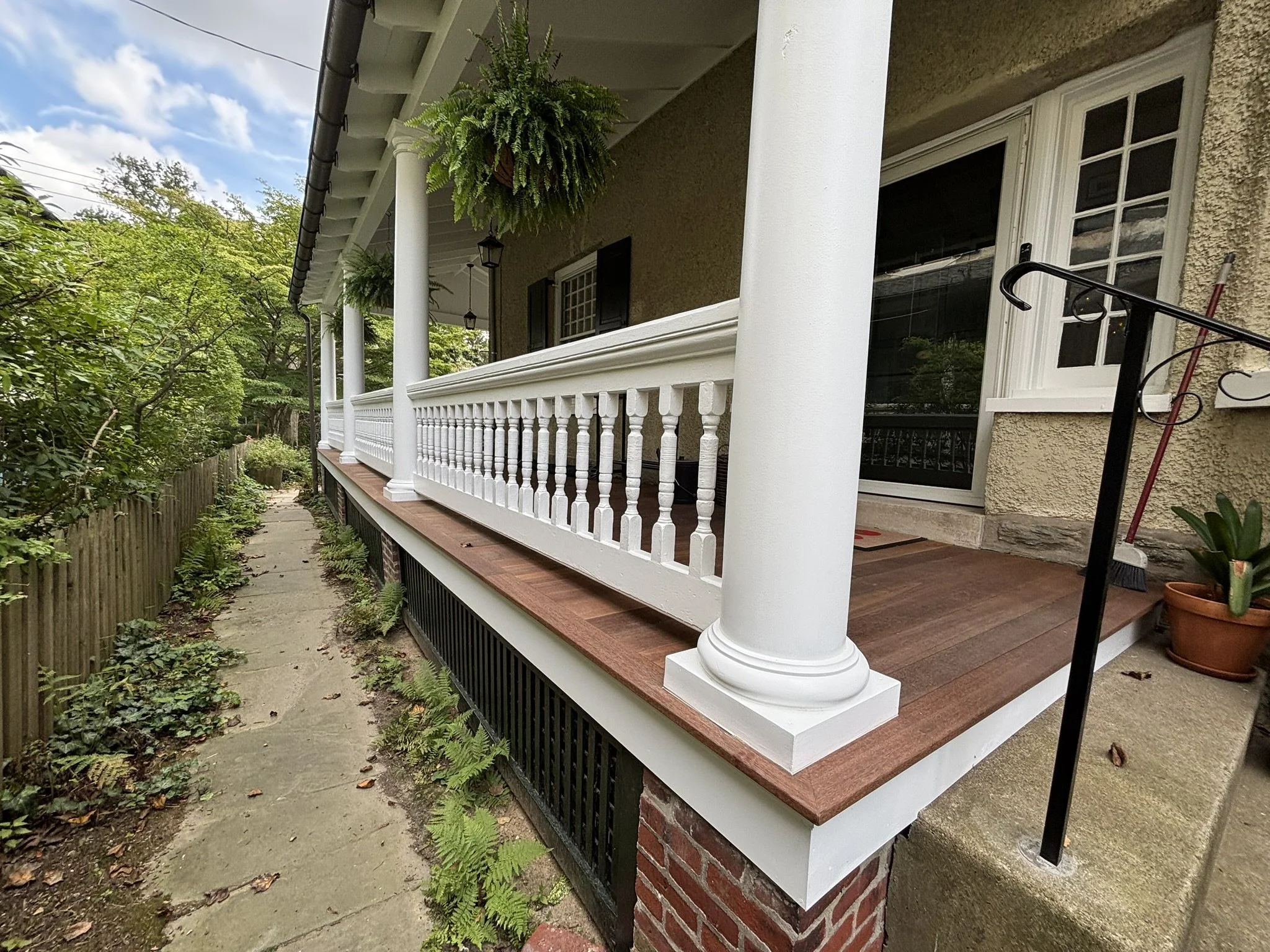 A house front porch with a white railing, hanging plants, and potted plants, with a concrete walkway and garden plants along the side.