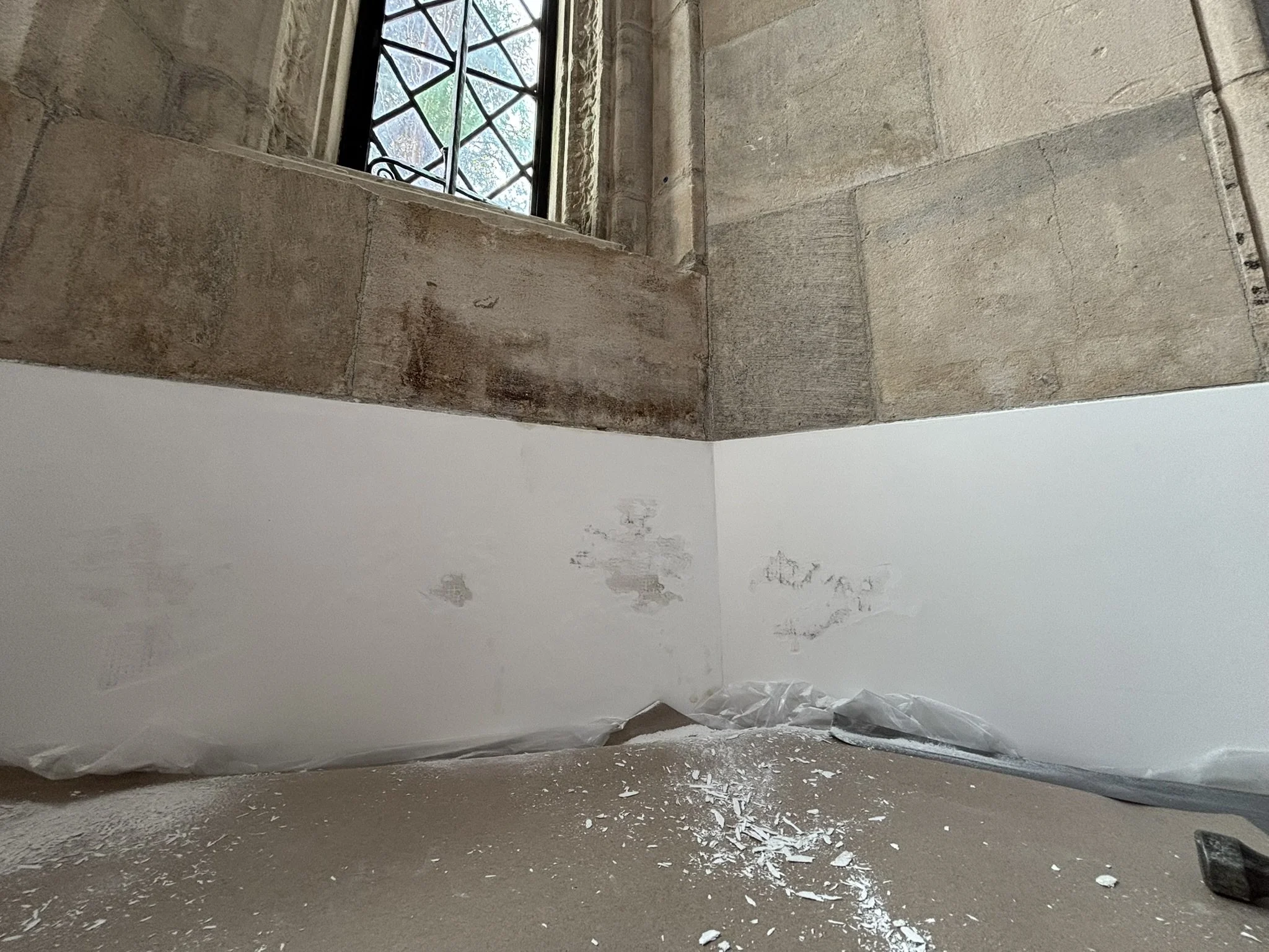 A corner of a room under renovation with a window, partially painted wall, and debris on the floor.