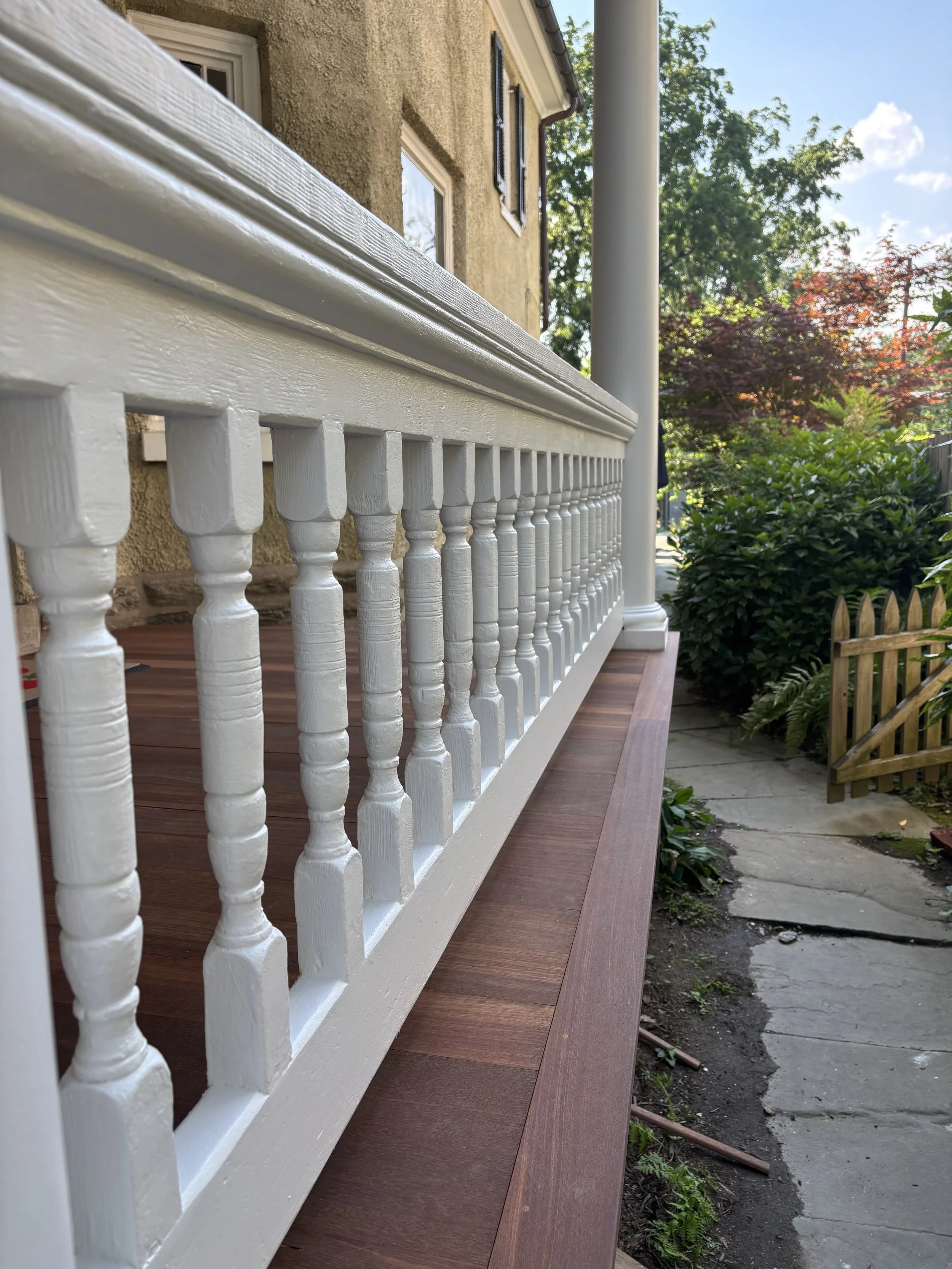 Close-up of a white wooden railing on a house porch, with a dirt and stone pathway, greenery, and a partly cloudy sky in the background.