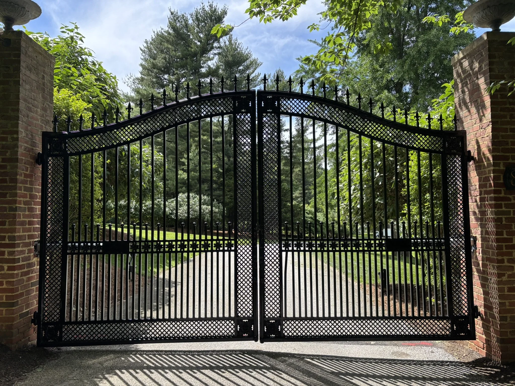 Black wrought iron gate between brick pillars with greenery and trees behind, casting shadow on the ground.