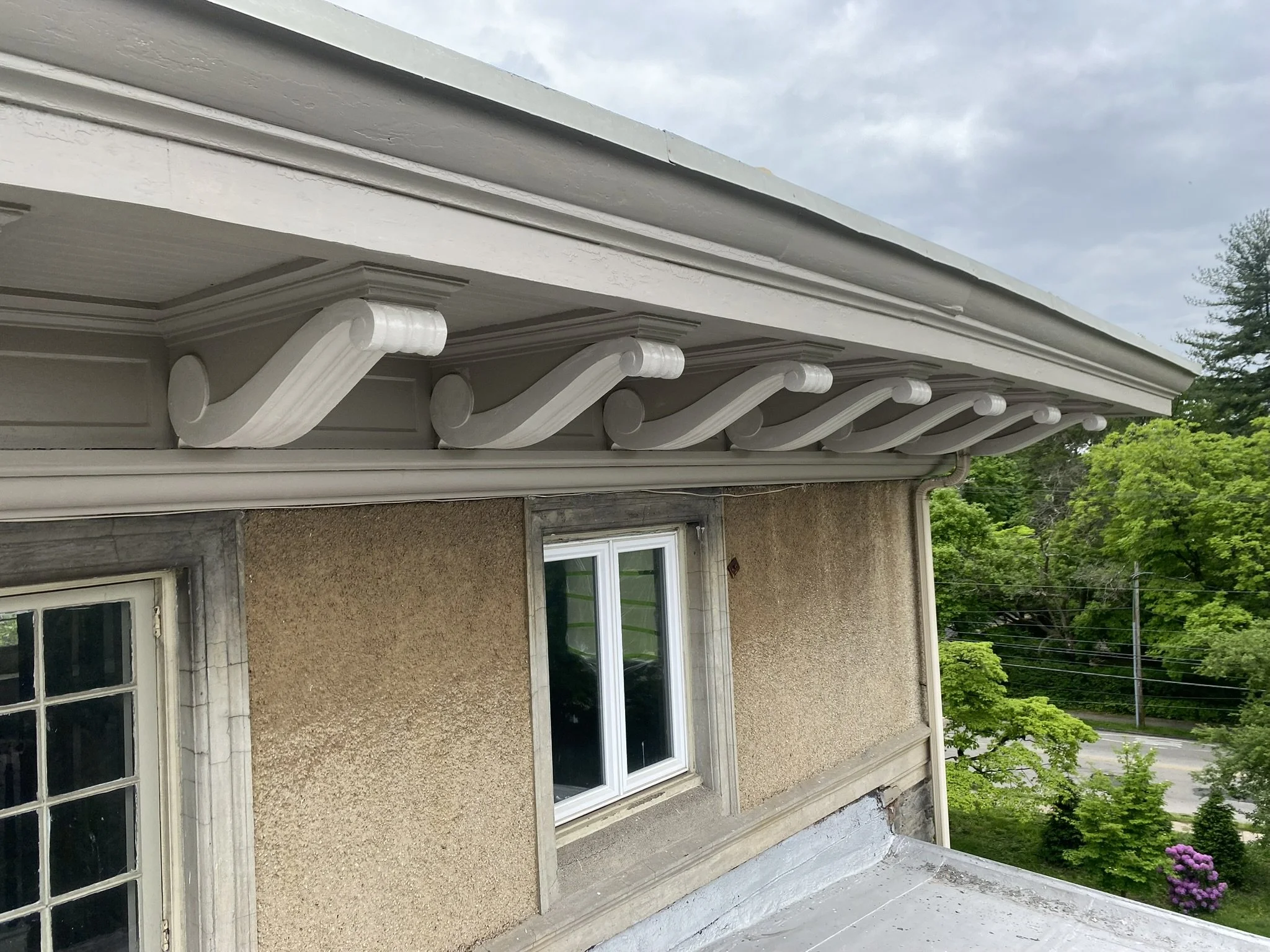 Upward view of a house's roof with decorative white roof brackets underneath the eaves, beige exterior wall with a window, green trees, and a partly cloudy sky.