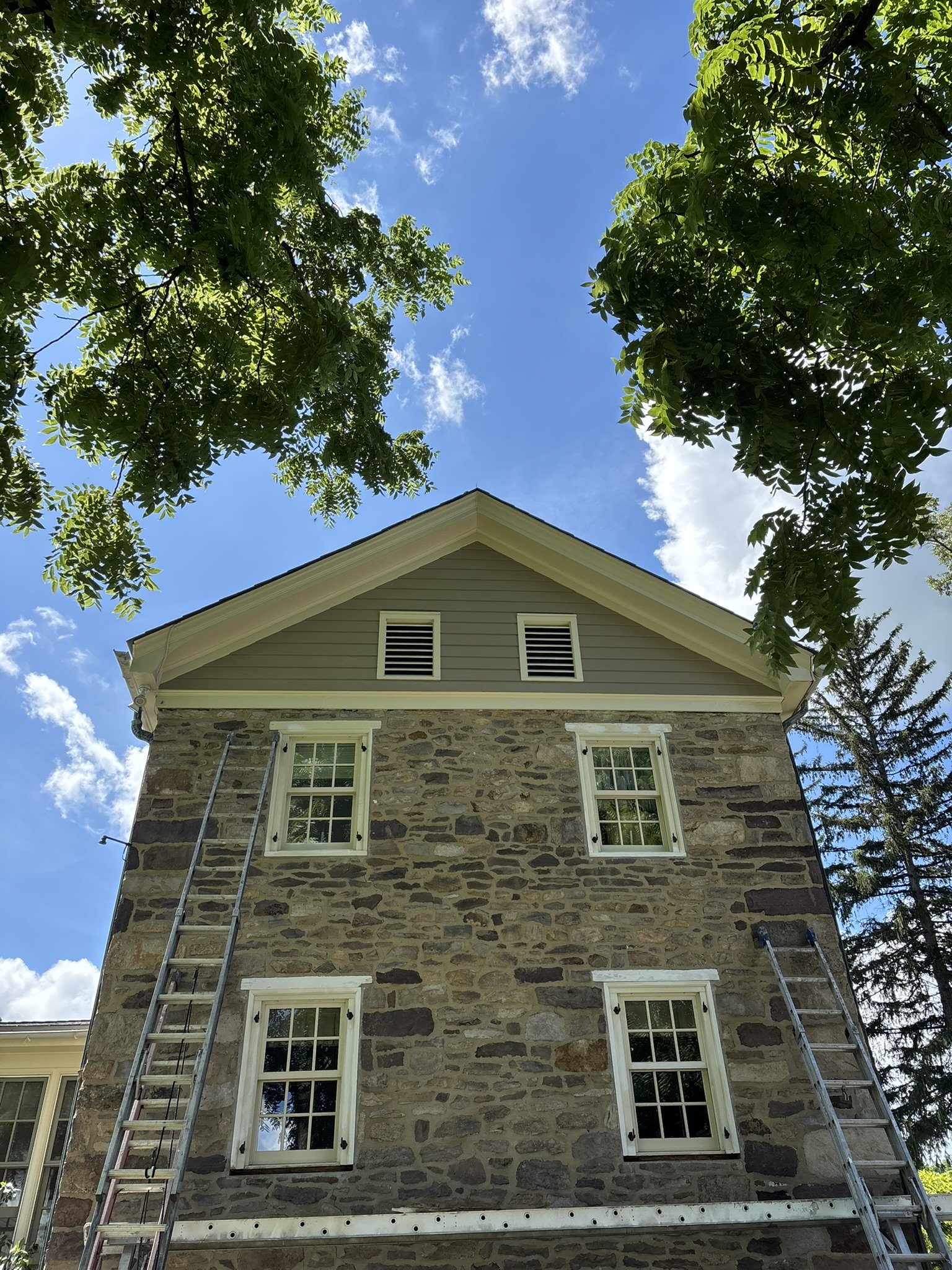 Looking up at a stone and siding house, with four windows, two on each floor, and two ladders leaning against it. Green tree branches frame the image, blue sky with clouds is visible above.