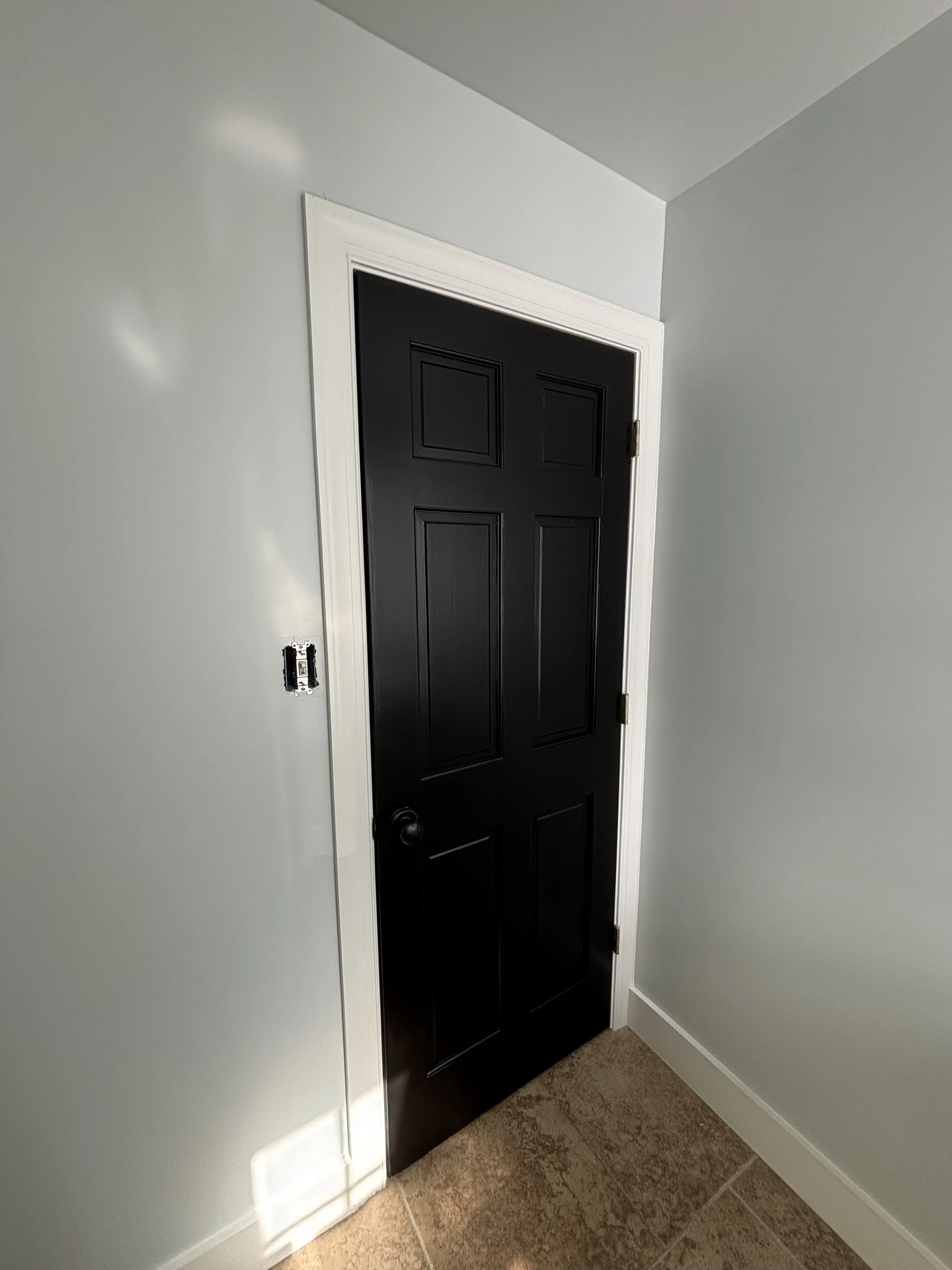 A black interior door with a bronze hinge, white trim, and a dark doorknob, set in a light-colored wall with a visible electrical outlet box without a cover.
