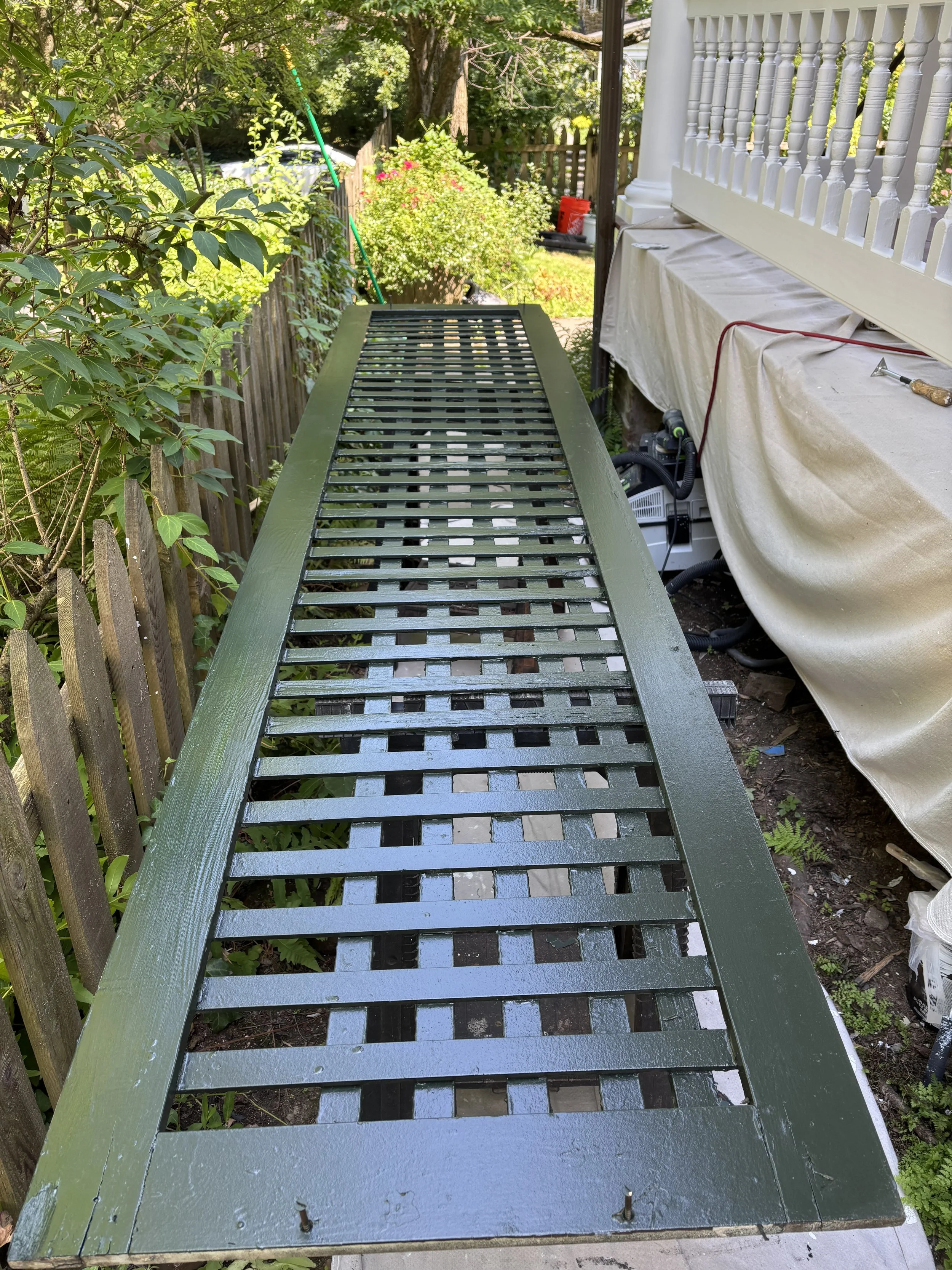 A freshly painted green wooden deck railing with slats, outdoors next to a house with a white porch railing, surrounded by plants and a wooden fence.