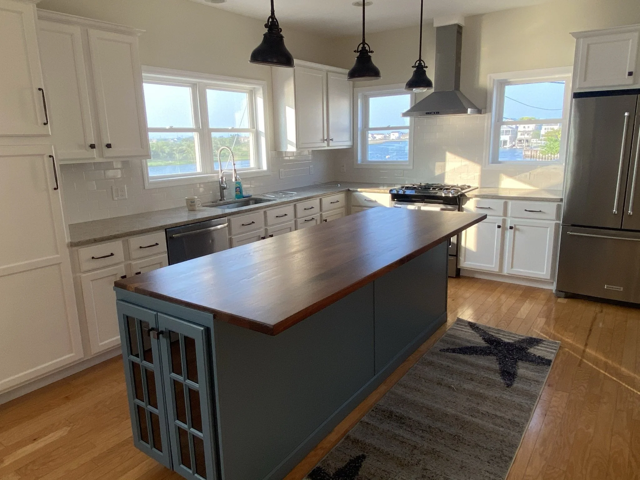 Modern kitchen with white cabinets, a wooden central island, stainless steel appliances, a gray range hood, and three hanging black pendant lights. There are three windows letting in natural light, and a small starfish rug on the wooden floor.