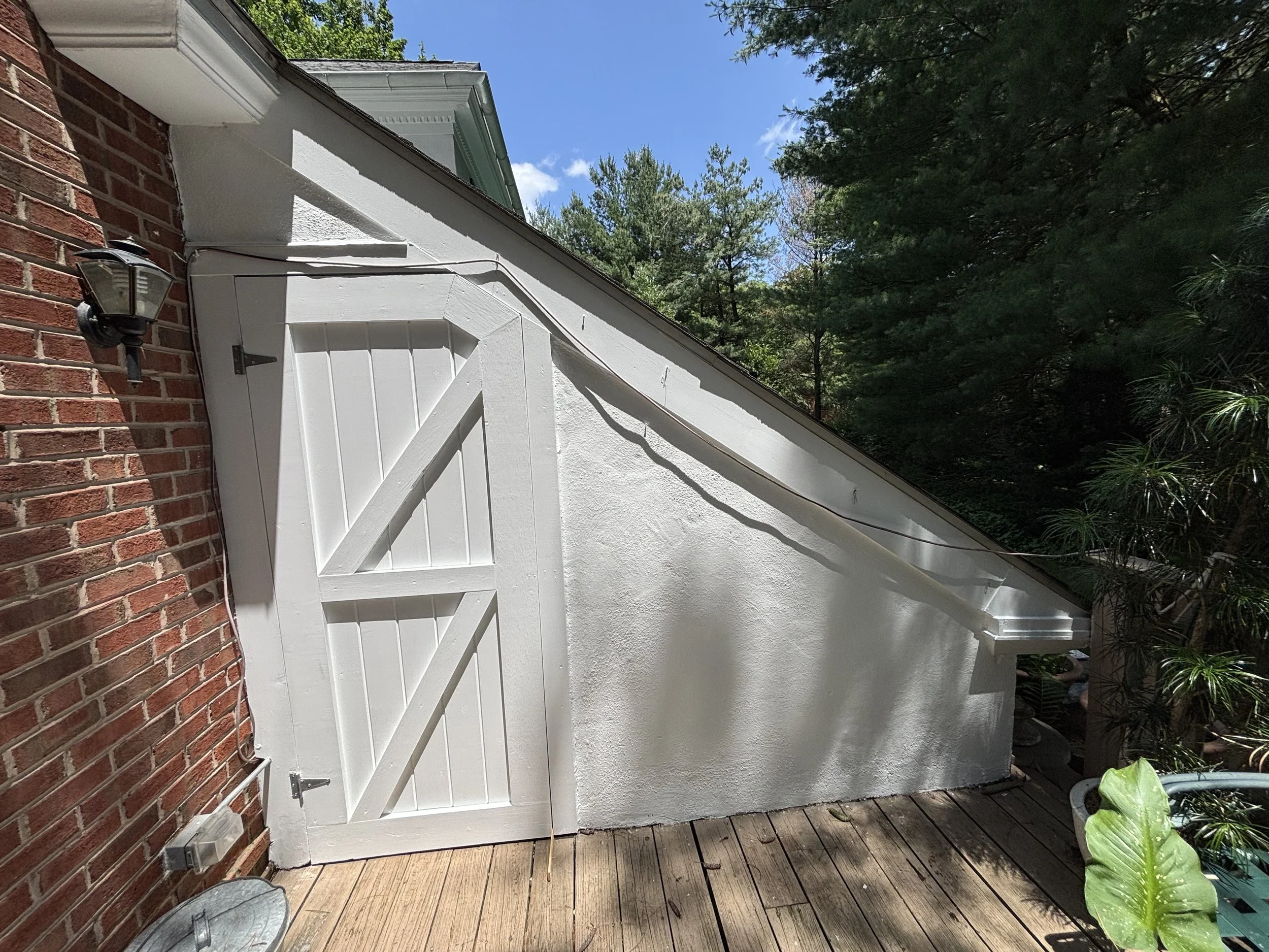 White shed with a sloped roof, attached to a brick wall, on a wooden deck, with green foliage and trees in the background, under a blue sky.